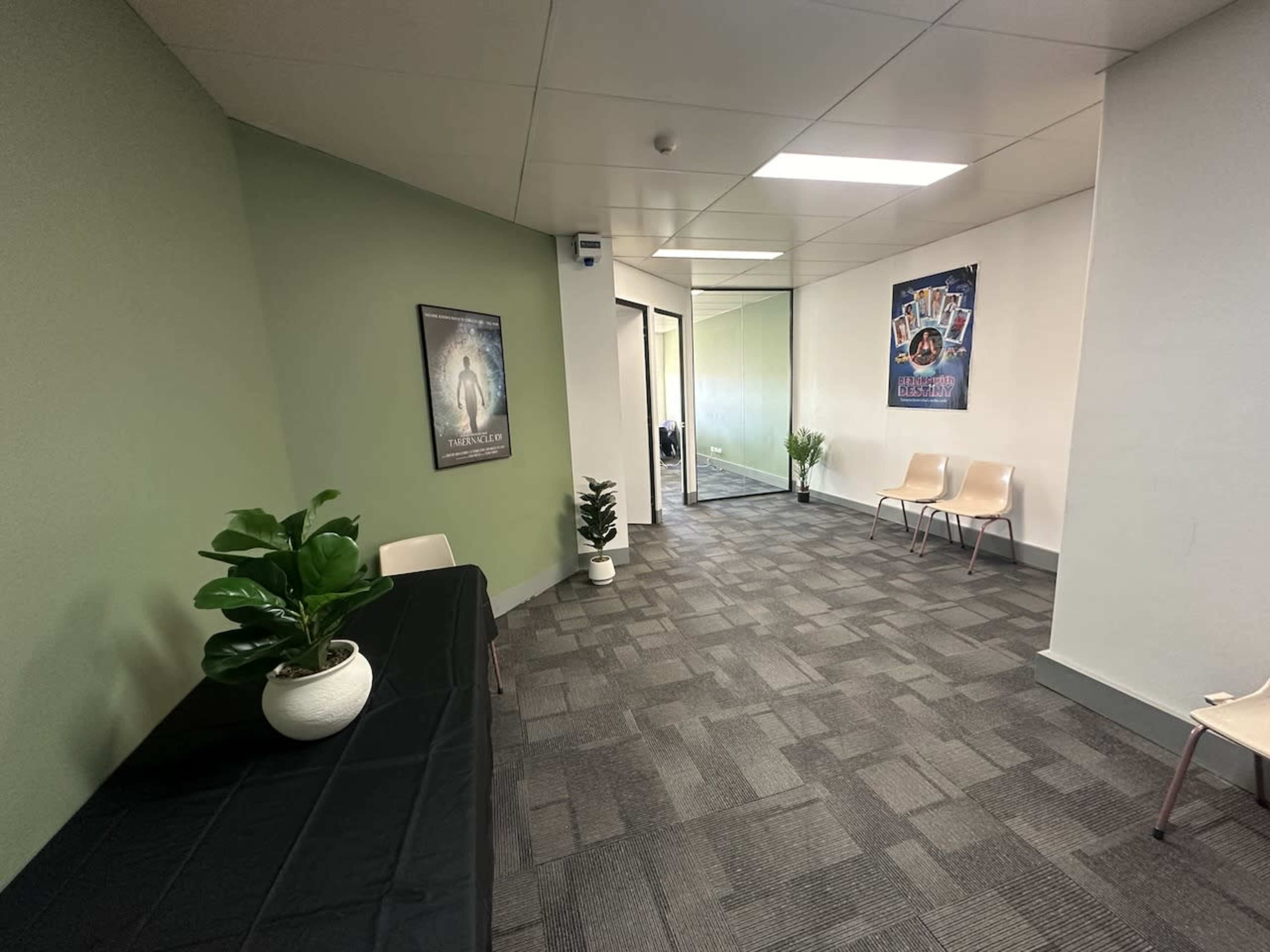 The image shows a corridor with a green wall, a table covered with a black cloth, two potted plants, and several chairs arranged along the wall.