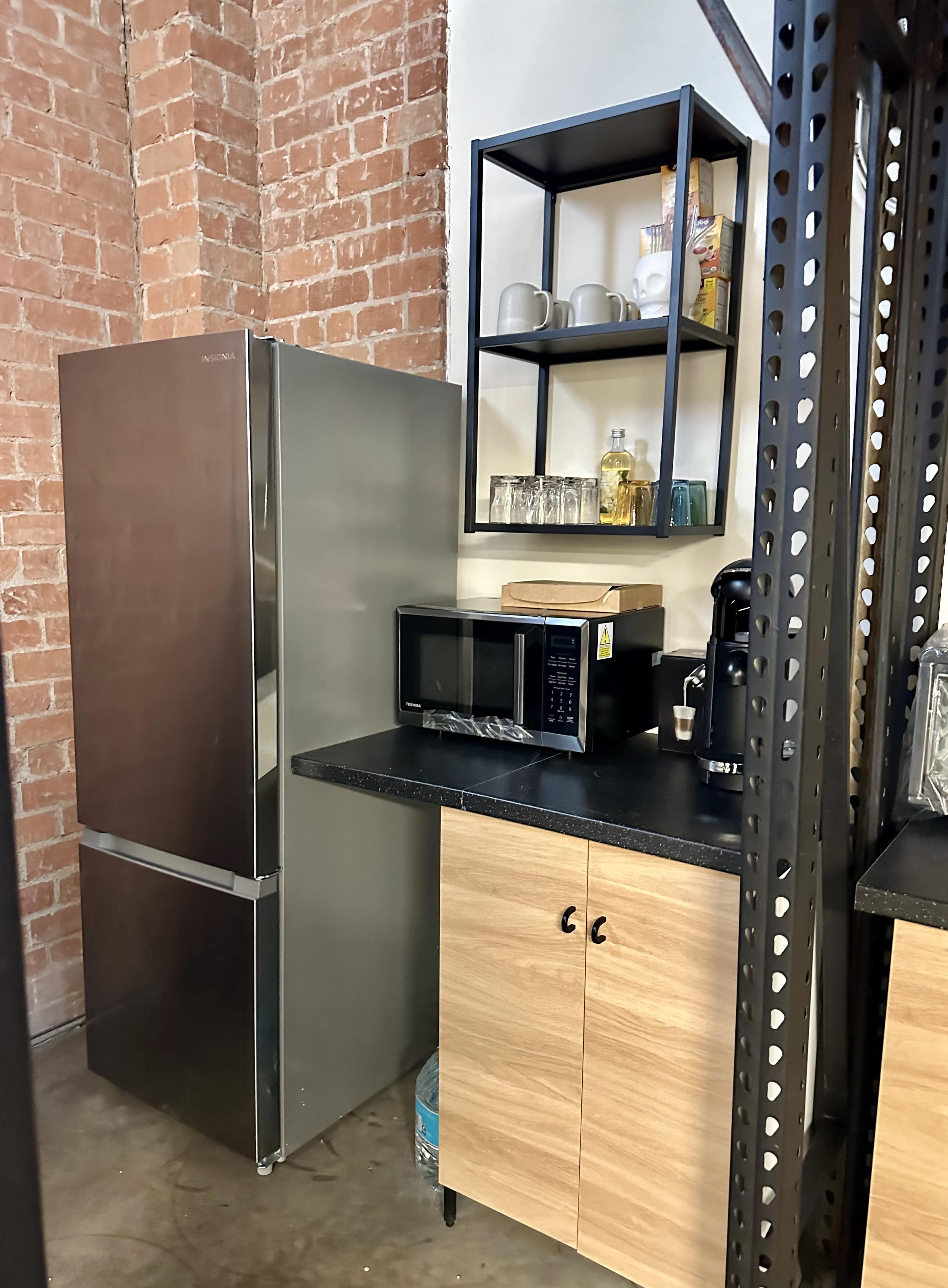 A kitchen area featuring a stainless steel refrigerator, a microwave on a black countertop, and a shelf holding various kitchen items against a backdrop of exposed brick walls.