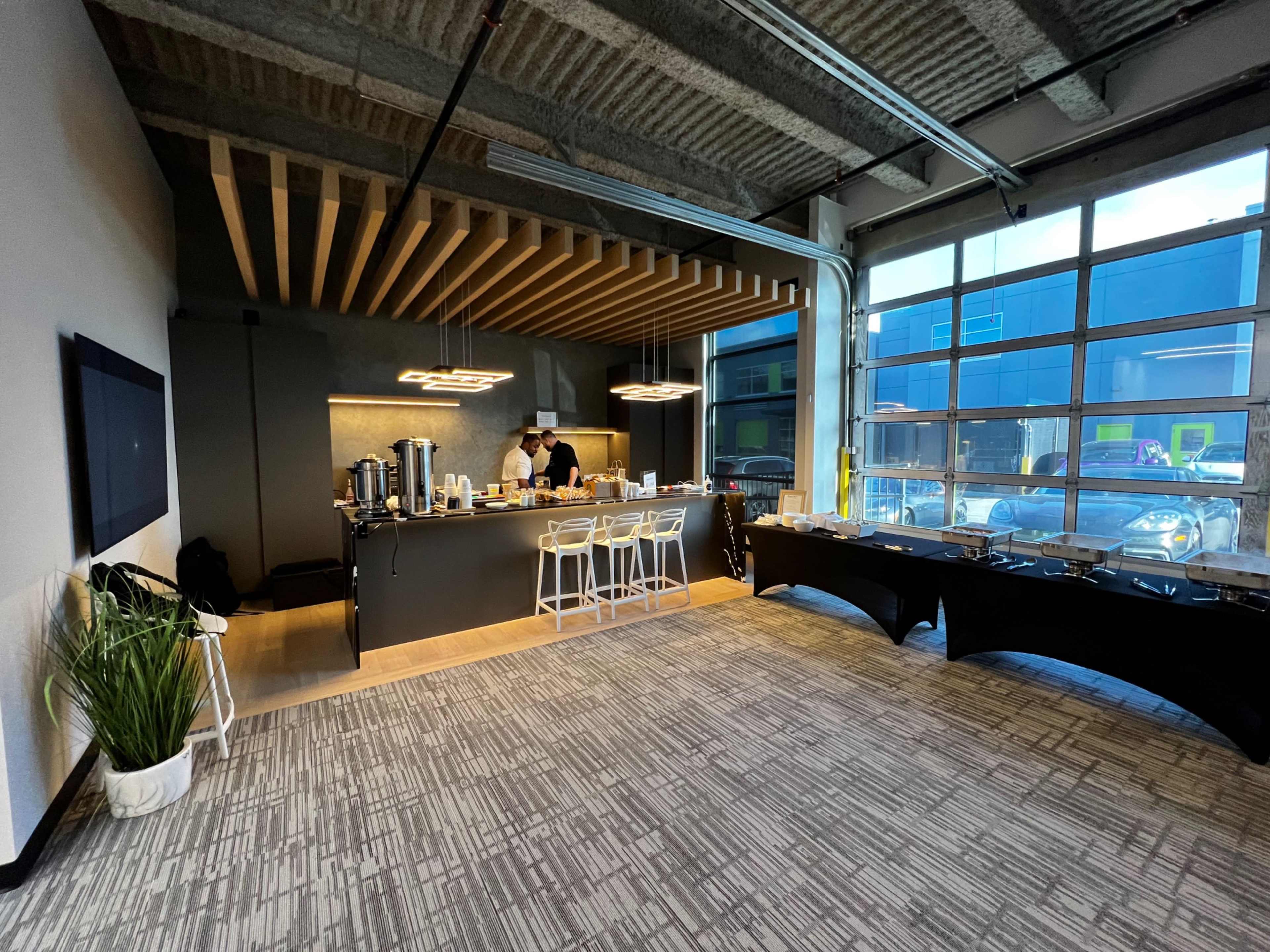 A modern indoor space features a dark countertop bar with a person preparing food, surrounded by chairs and a buffet table, with large windows allowing natural light.