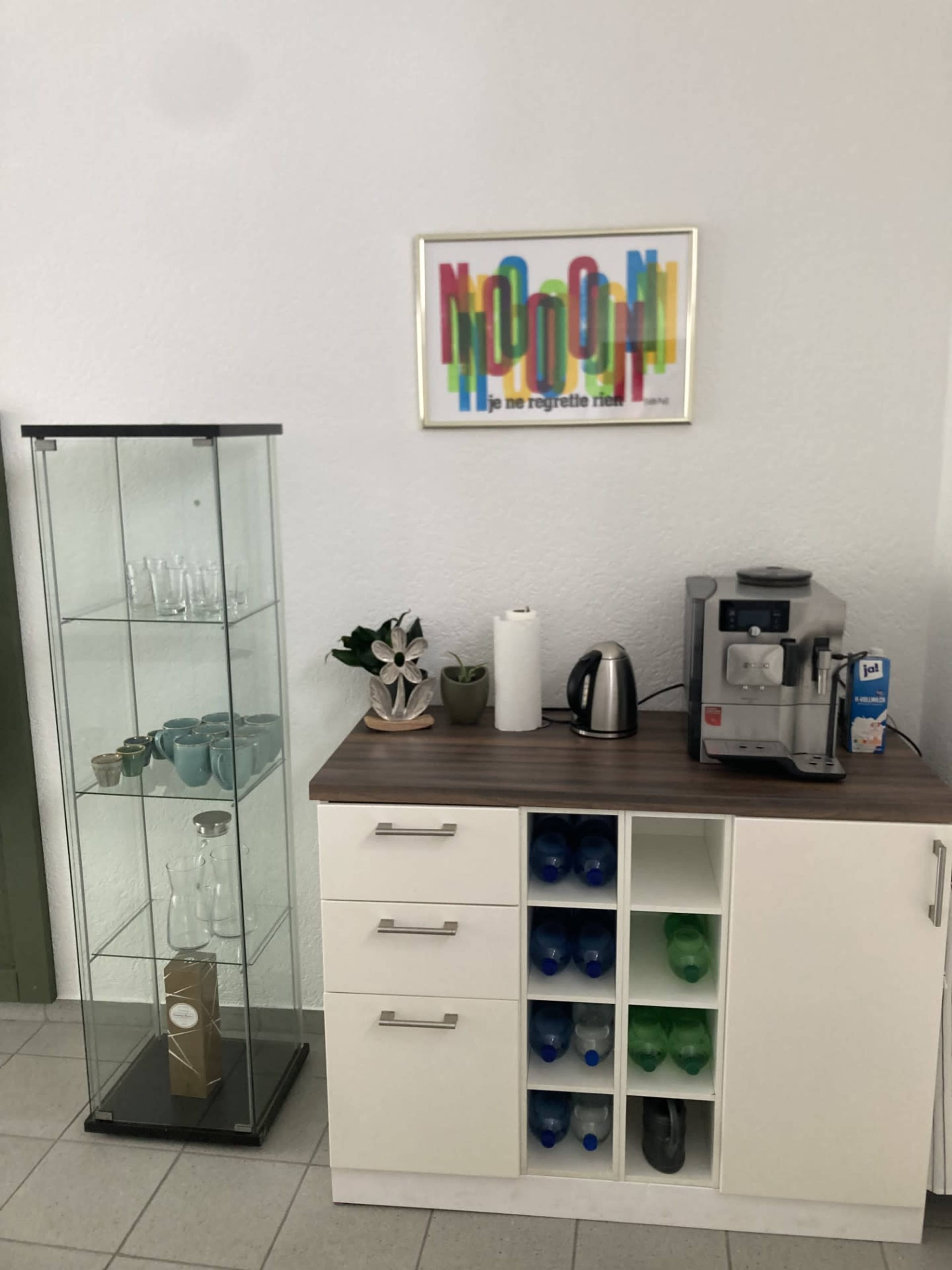 The image shows a modern kitchenette featuring a glass display cabinet, a coffee machine, a kettle, and a storage unit with visible water bottles.