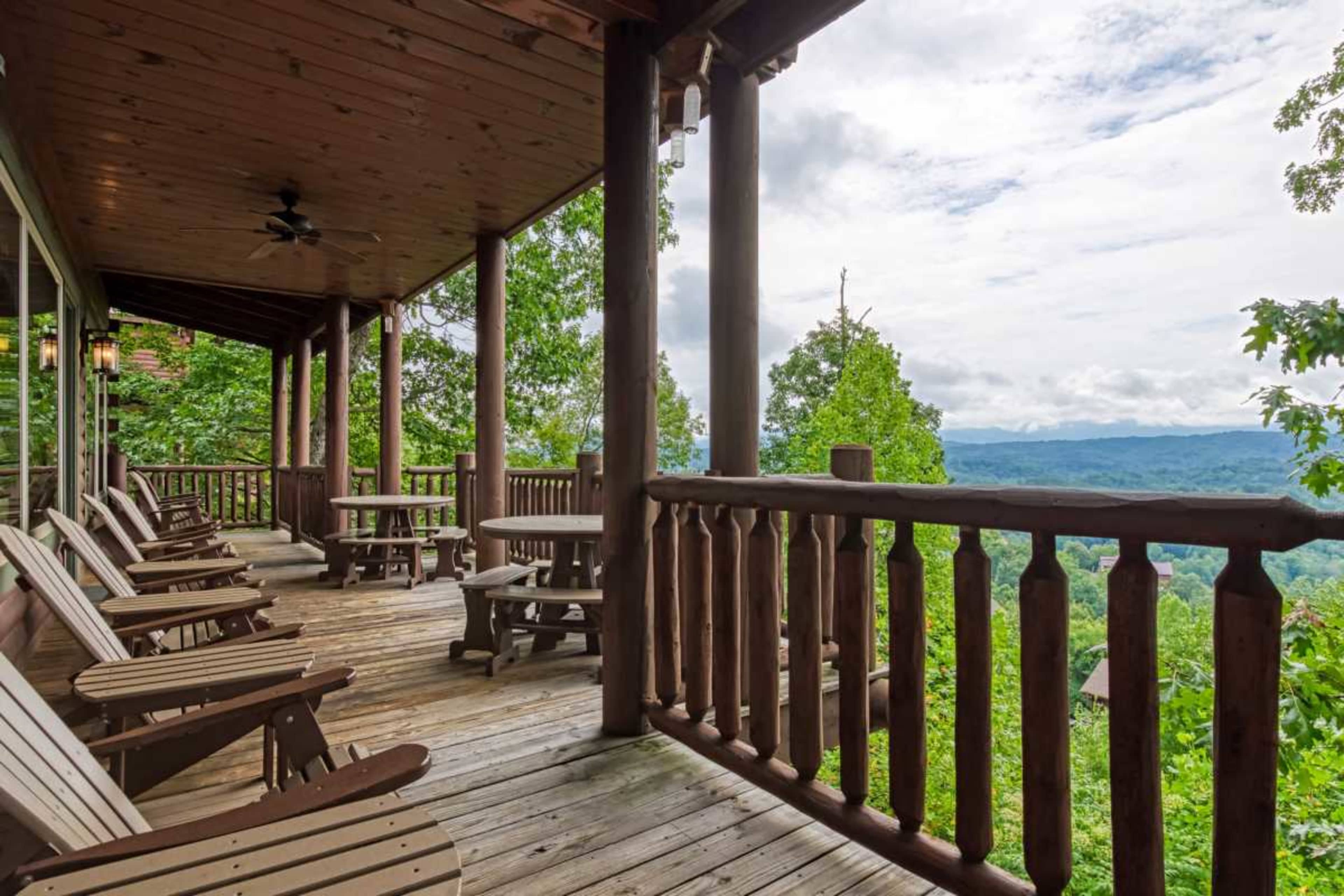 The image shows a wooden deck with several chairs and tables overlooking a scenic valley surrounded by trees and mountains under a cloudy sky.