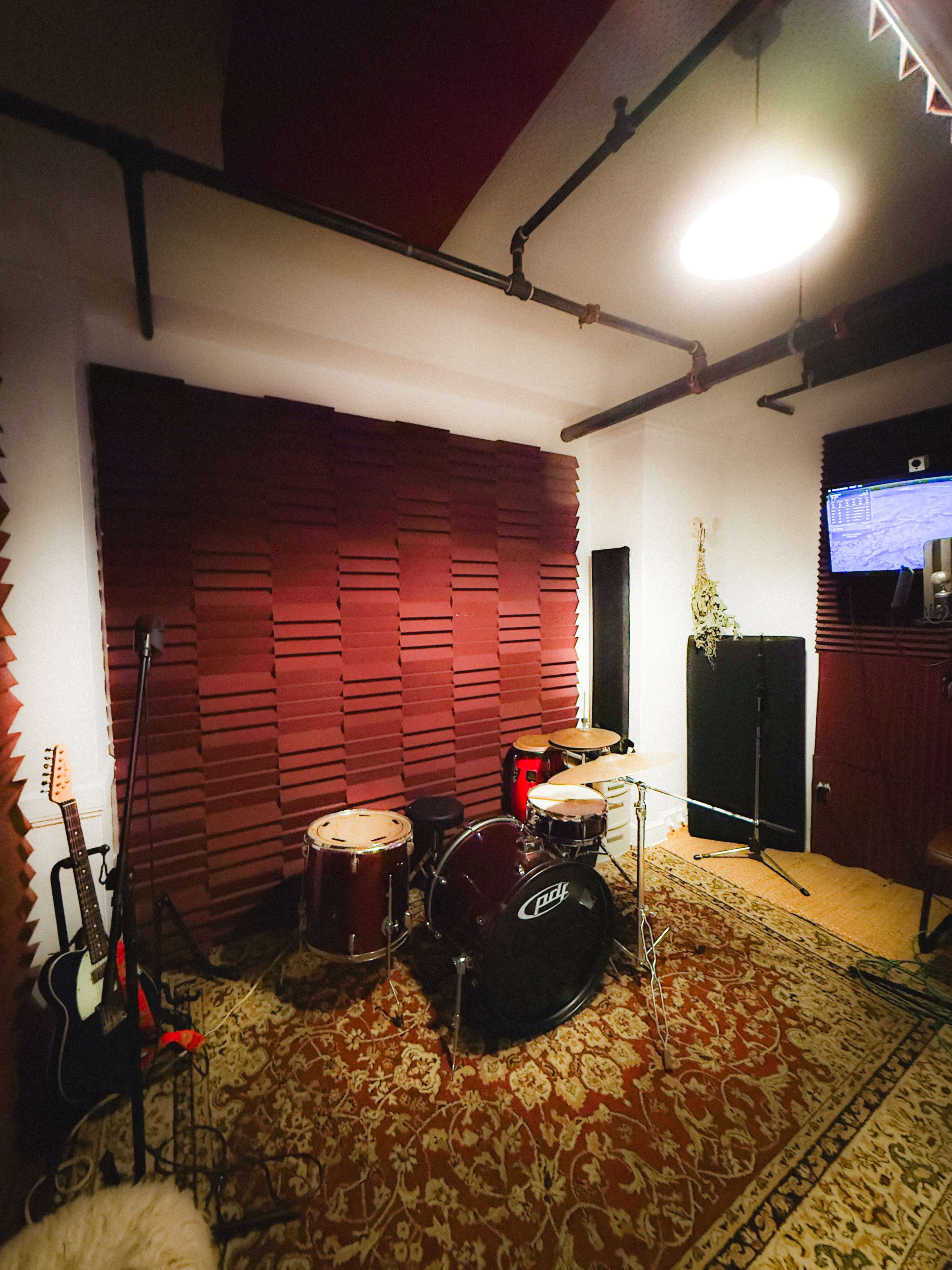 A music studio features a drum set, an electric guitar, and soundproofing panels on the walls.