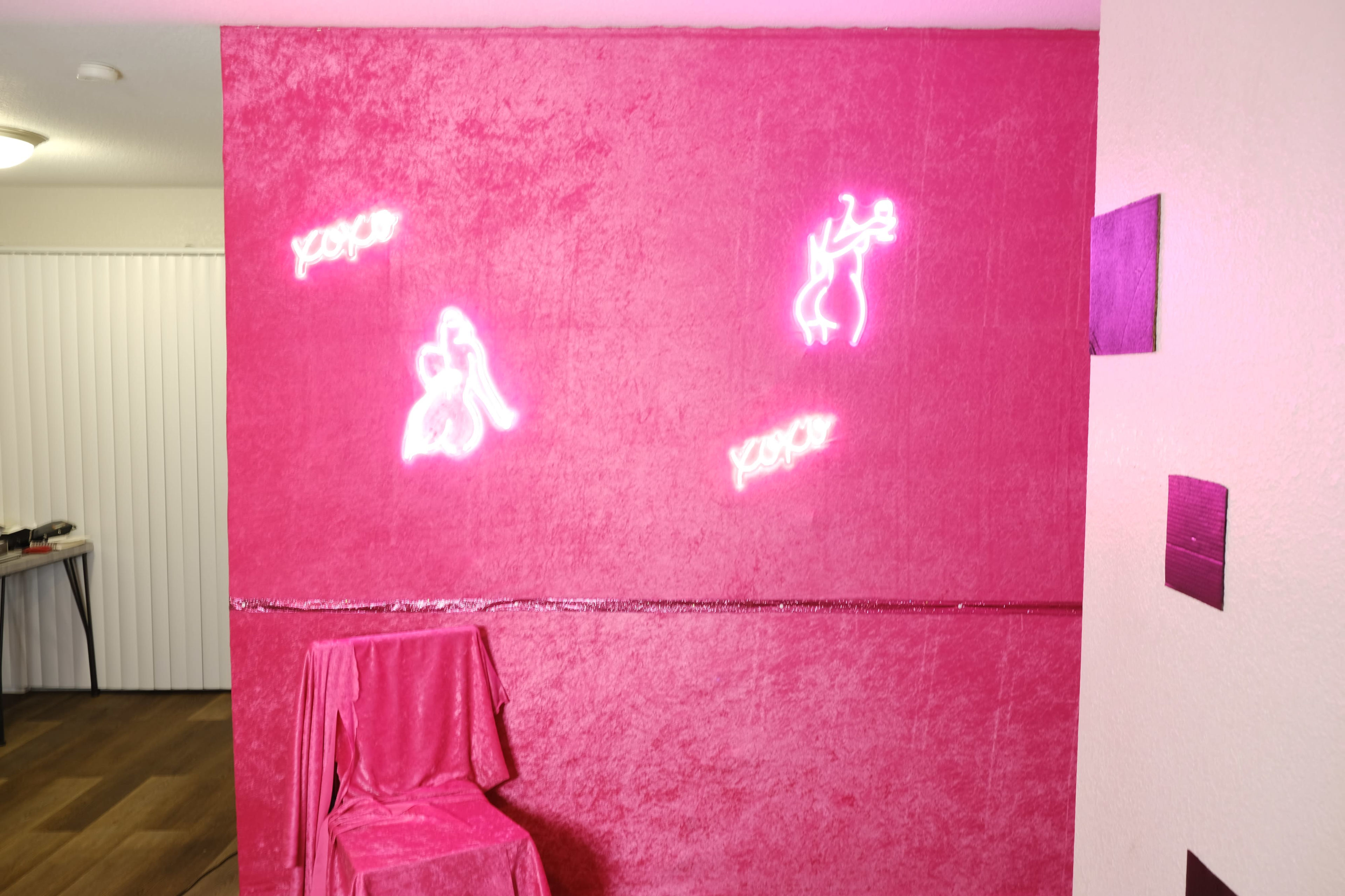 A pink textured wall featuring three neon signs and a covered chair in front of it.