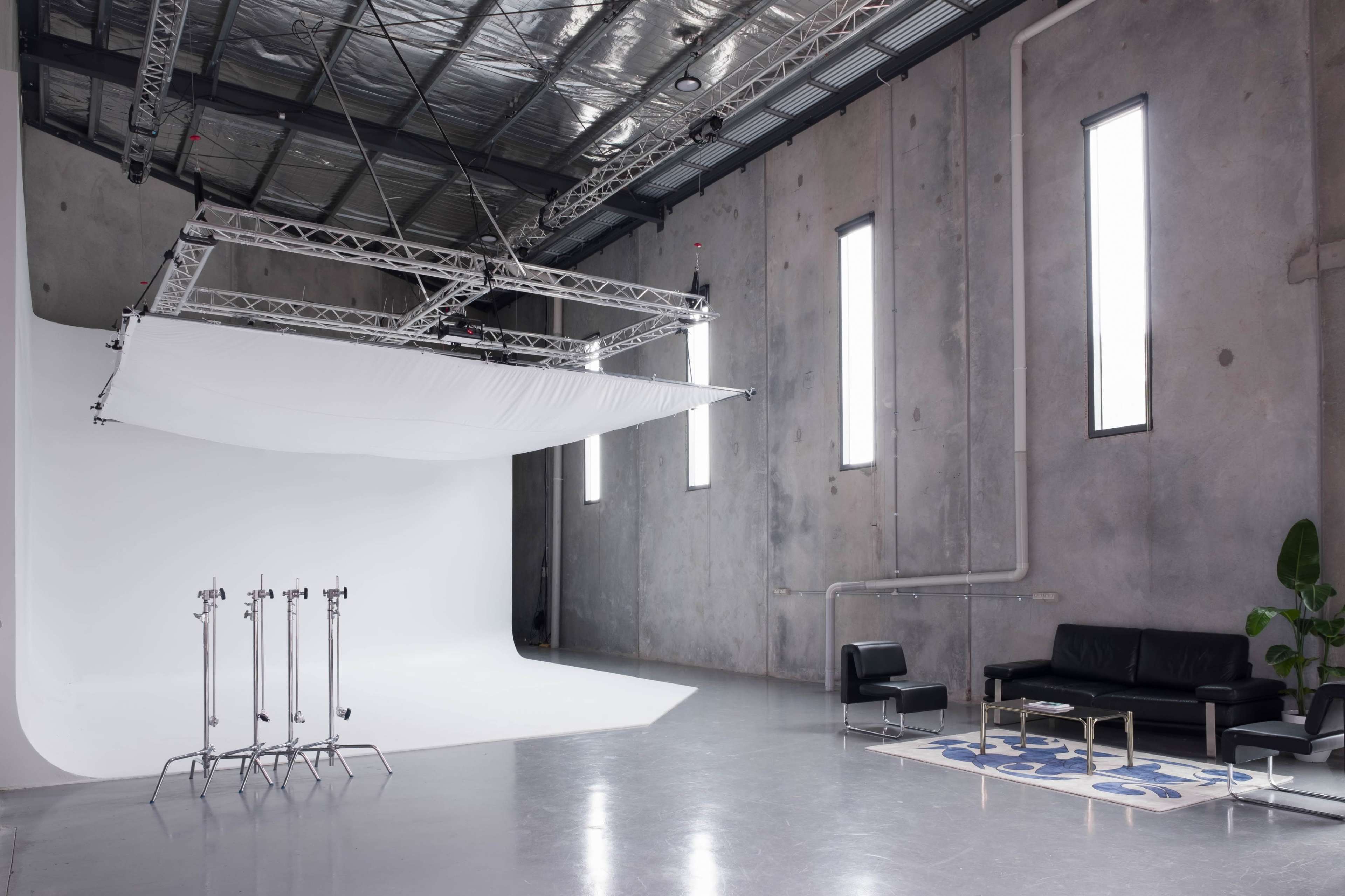 A modern photo studio with a white backdrop, studio lighting stands, and a minimalist black sofa set against a concrete wall.