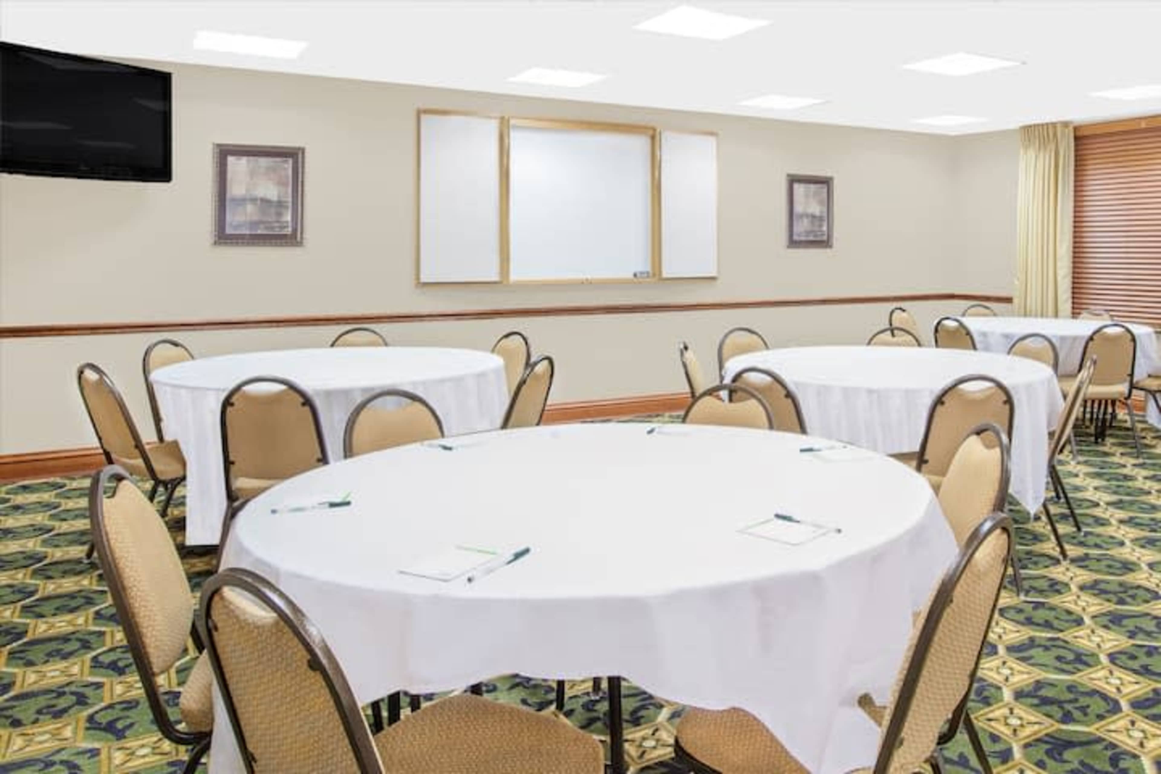 A room set up for a meeting, featuring round tables covered with white tablecloths and chairs arranged around them, with a blank whiteboard and a television visible on the wall.