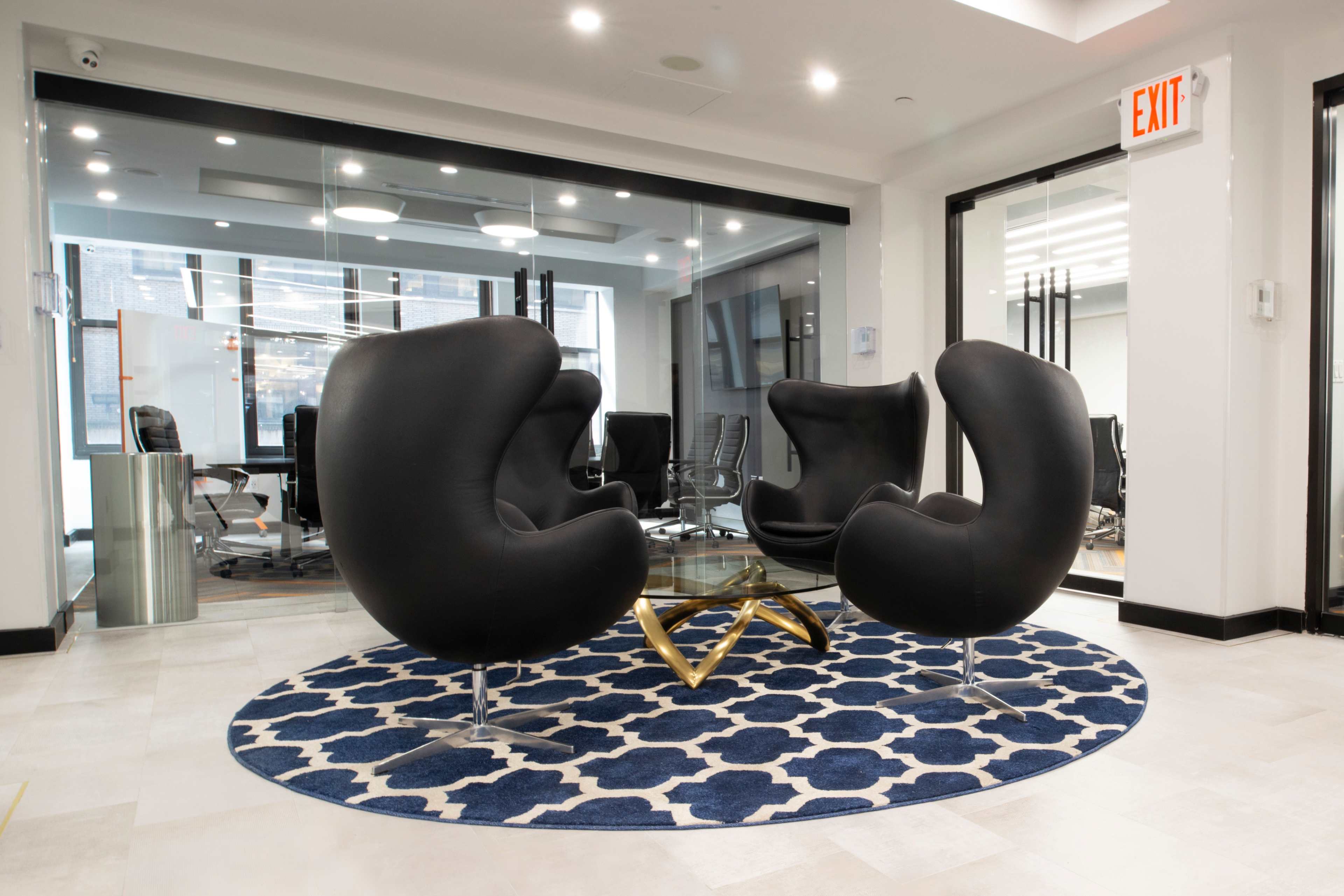 The image shows a modern office space featuring two black curved chairs surrounding a glass coffee table on a patterned blue rug.