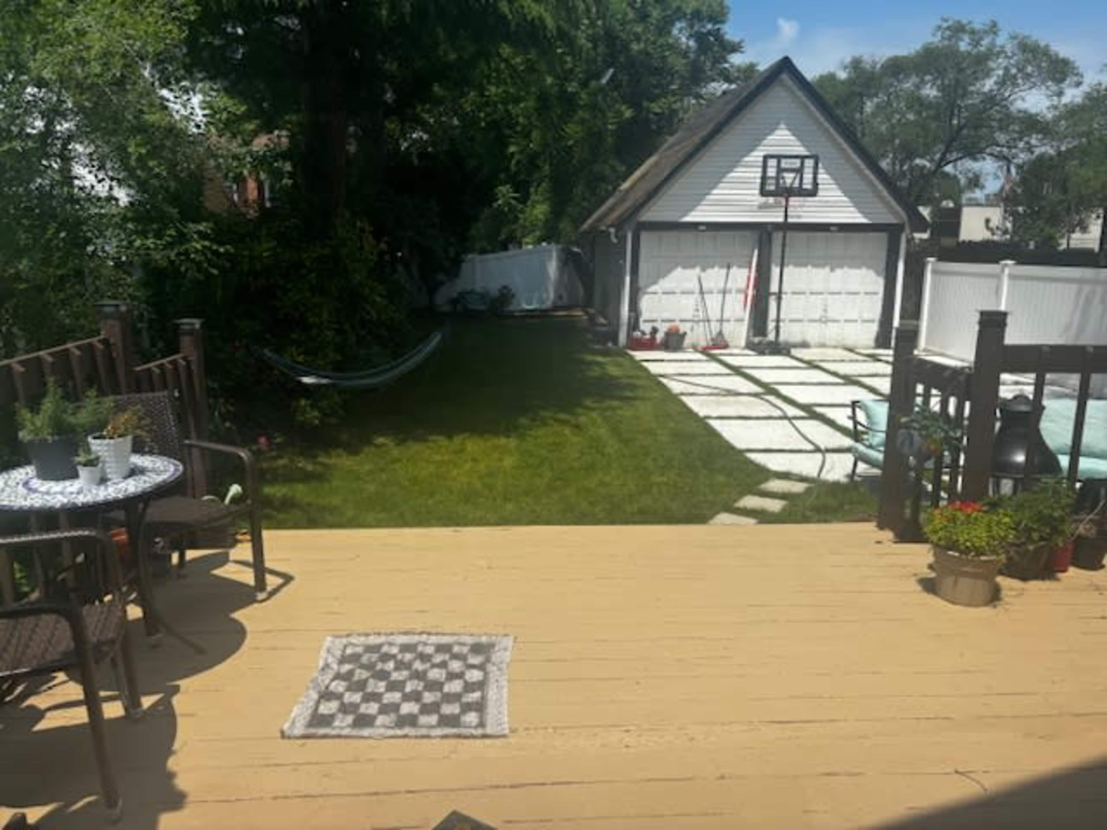 A backyard with a wooden deck, two chairs and a small table, a grassy area, a hammock, a pathway of pavers, a garage with basketball hoop, and a glimpse of a pool.