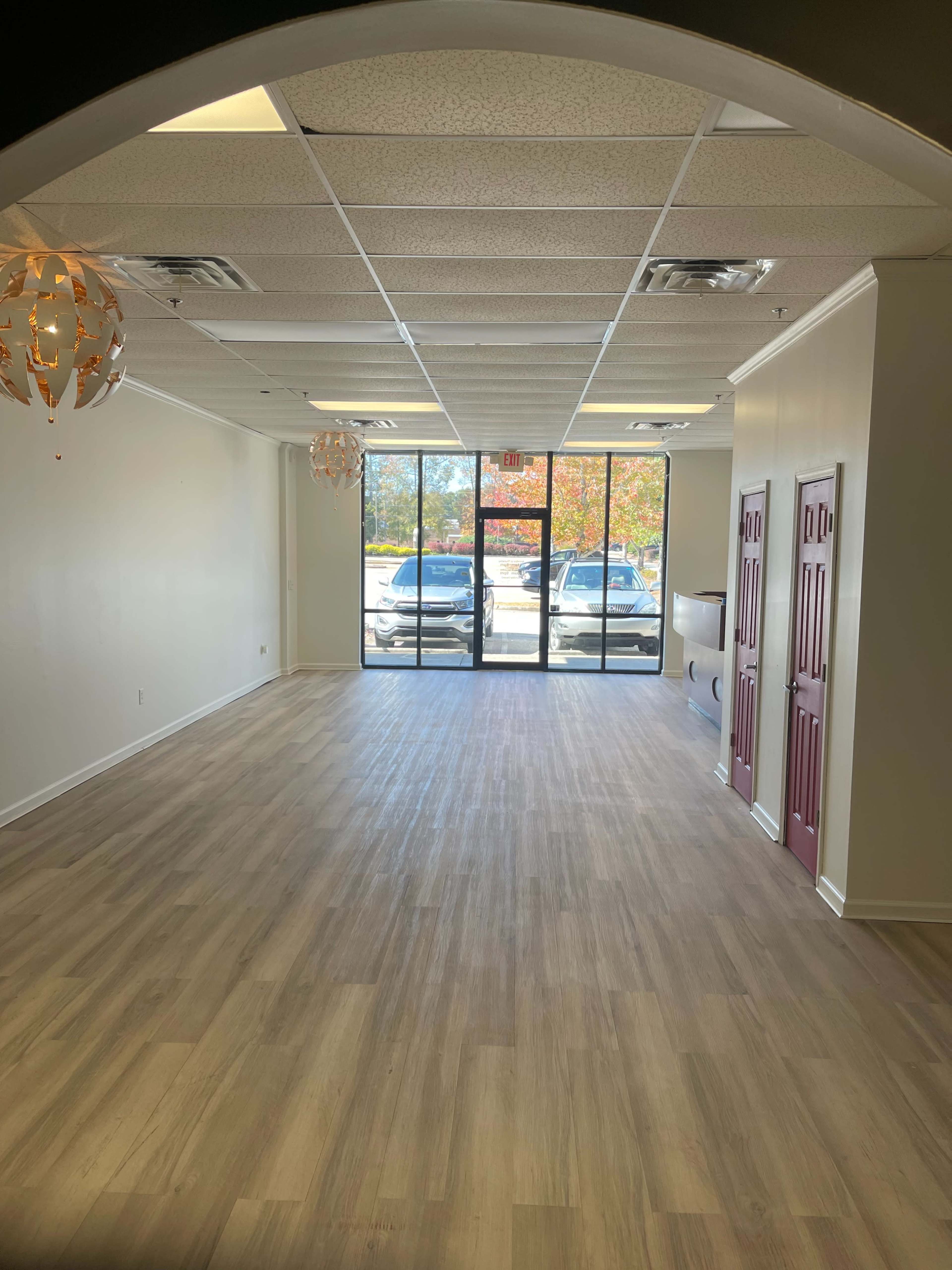 An empty commercial space with light-colored walls, wooden flooring, and large front windows allowing natural light.