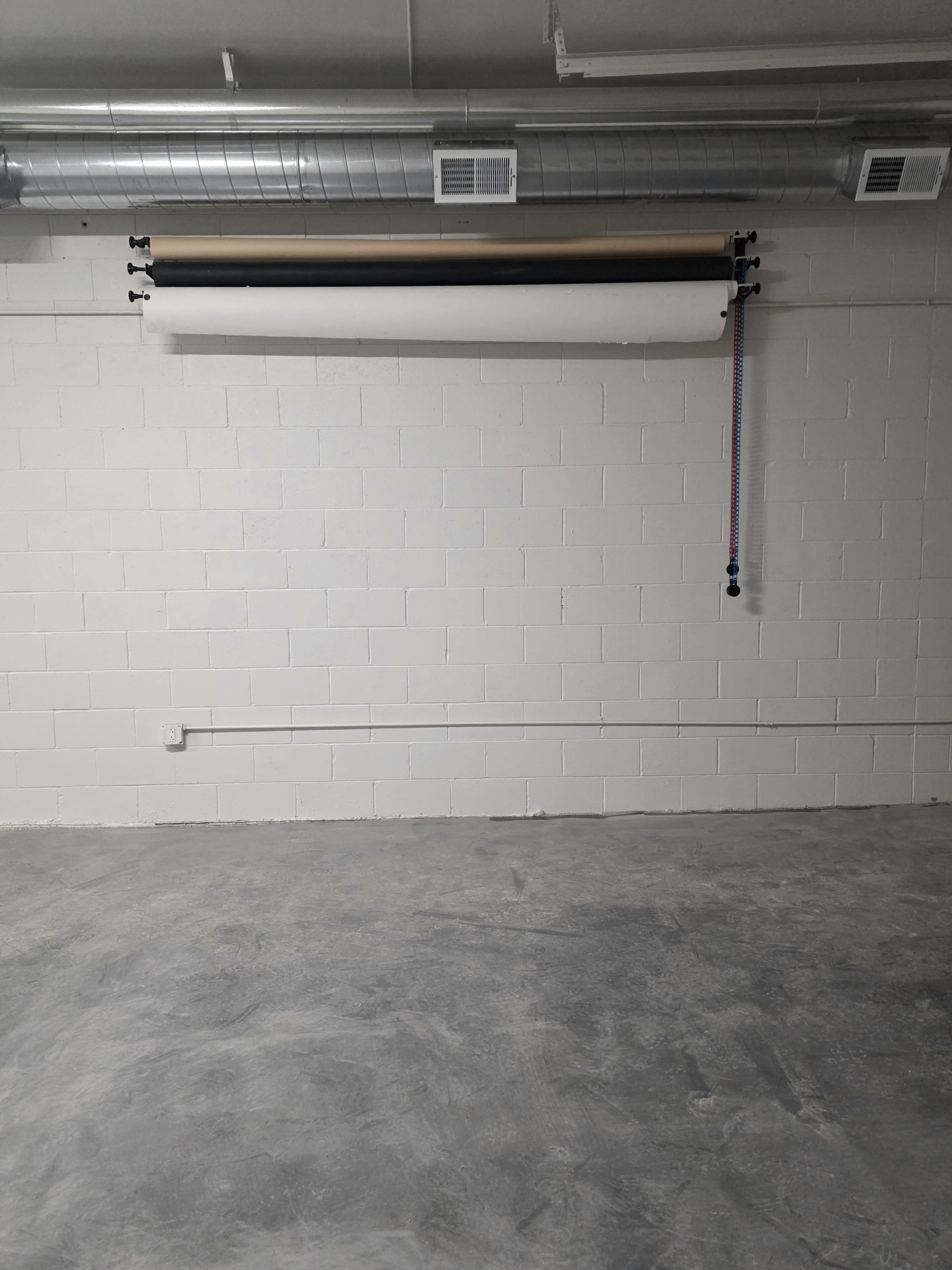 The image shows a bare wall with a roll of backdrop paper hanging from a mounted rail, and a few electrical outlets on the concrete floor beneath.