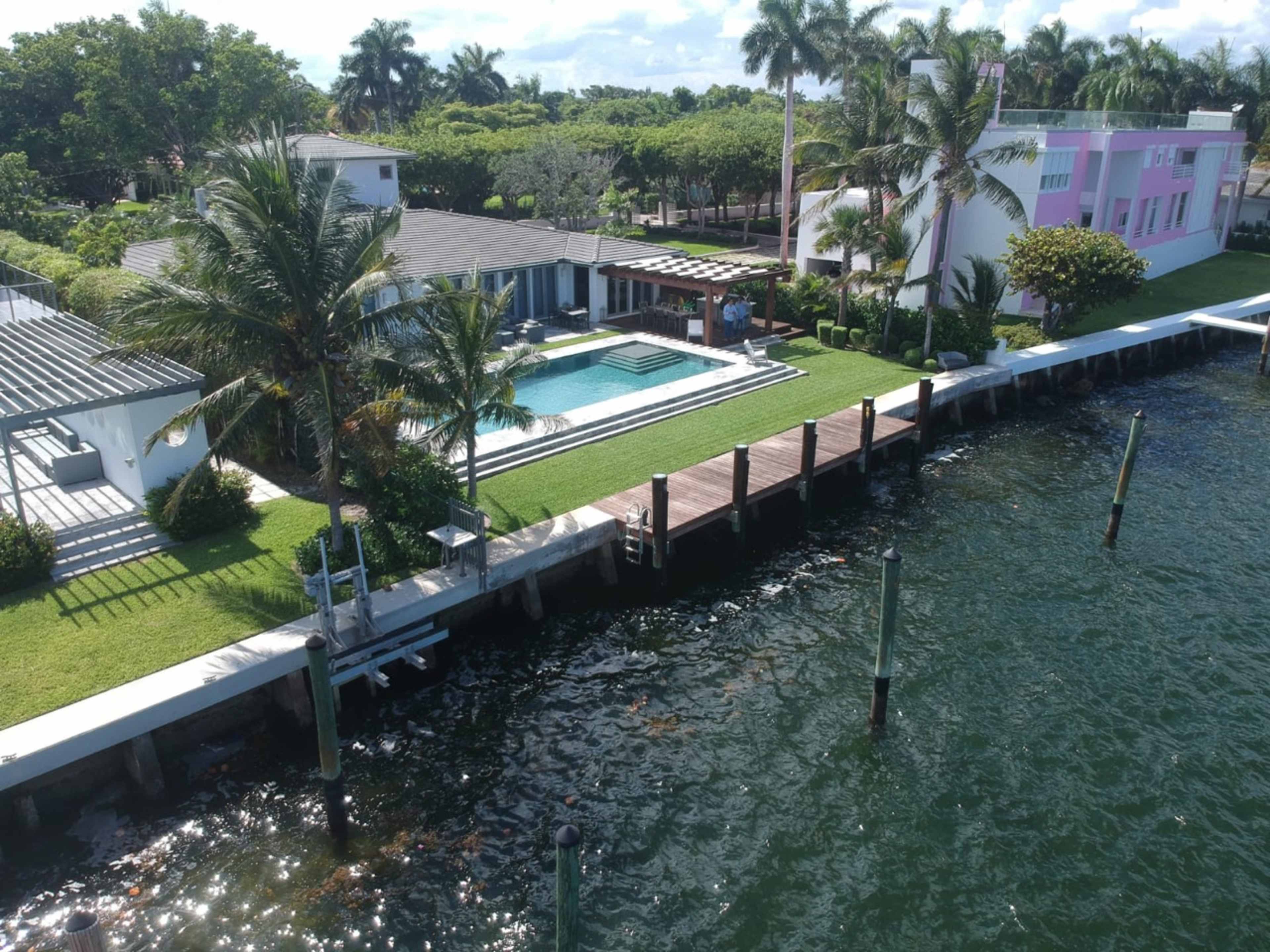 The image shows a waterfront property featuring a pool, palm trees, and a dock on a canal surrounded by green lawns and residential buildings.