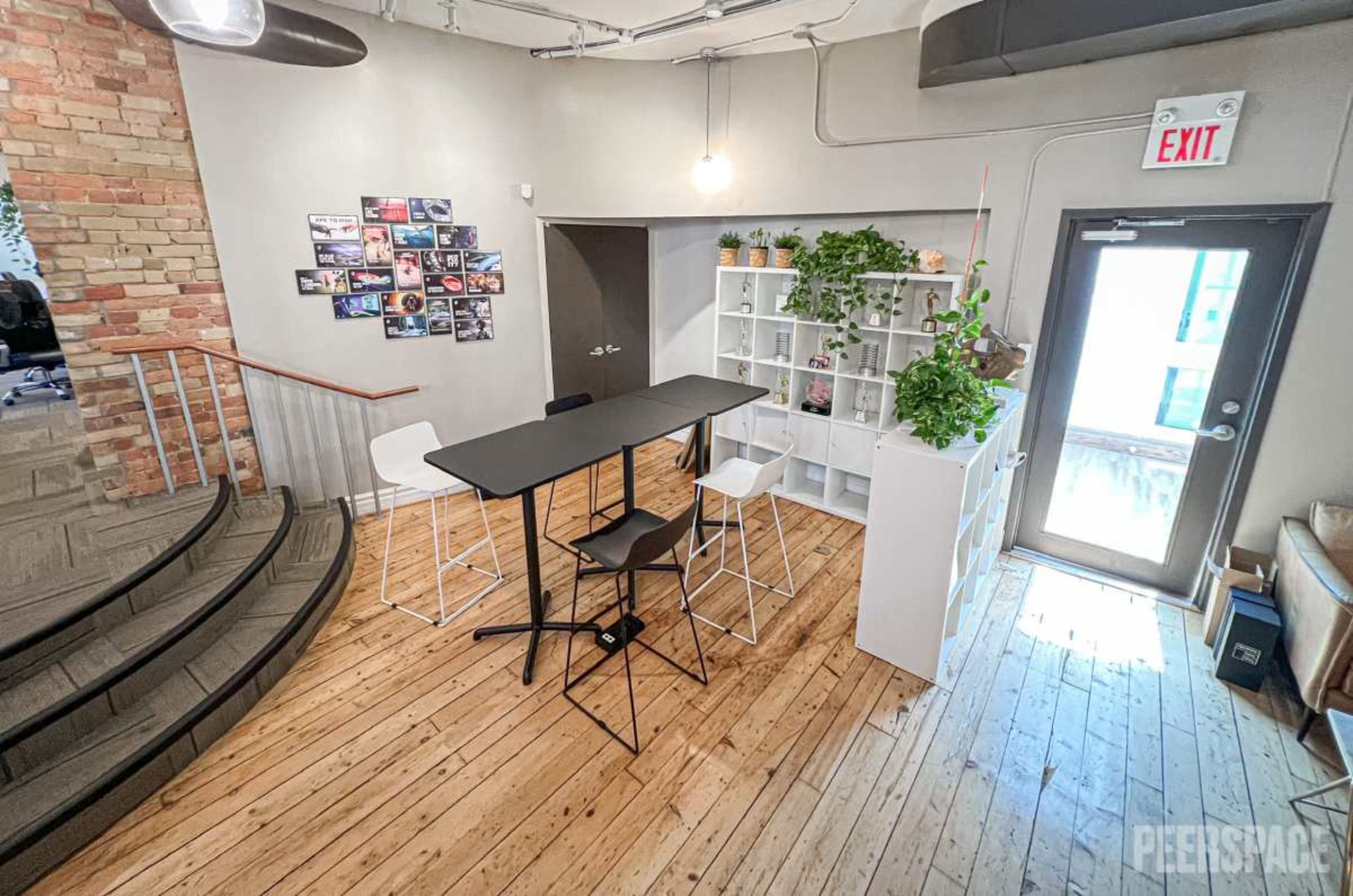 The image shows a modern workspace featuring a tall table with chairs, surrounded by plants and a gallery wall of photos.