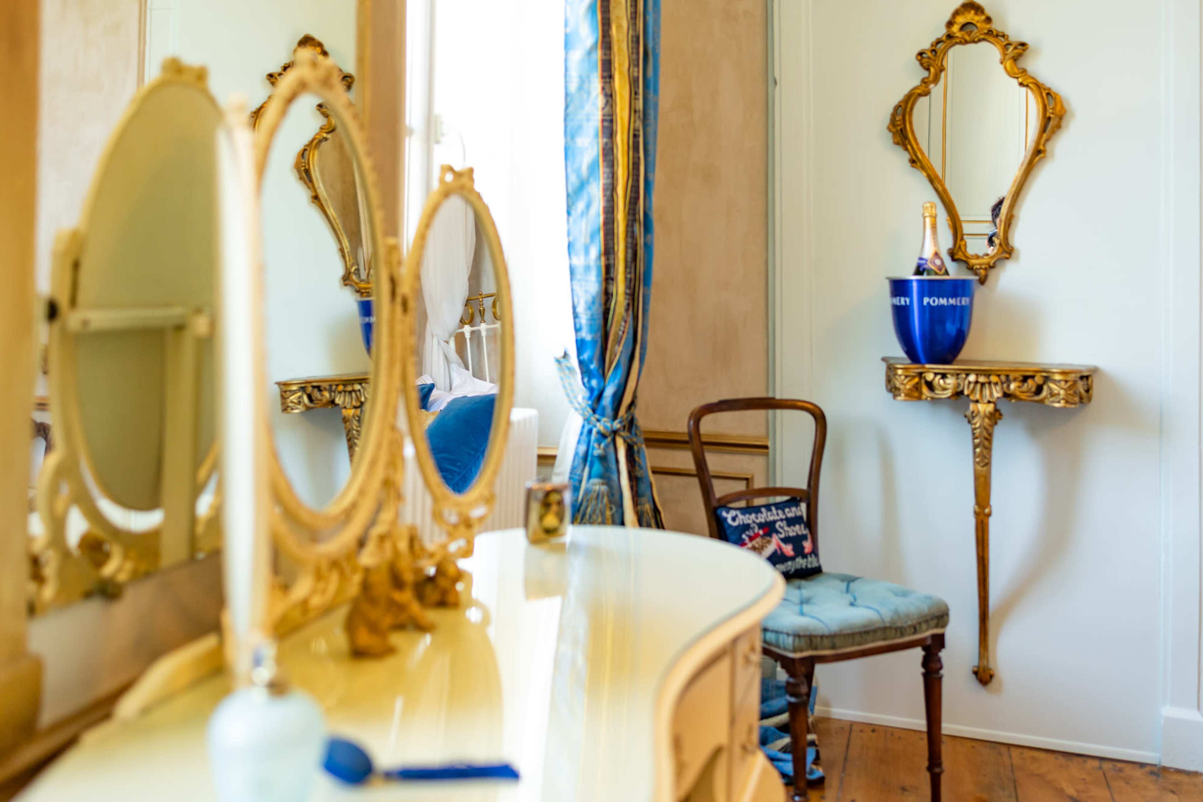 A vintage dressing table with mirrors and a gold-framed wall shelf holding a champagne bottle is set against a light-colored wall in a room decorated with blue curtains.