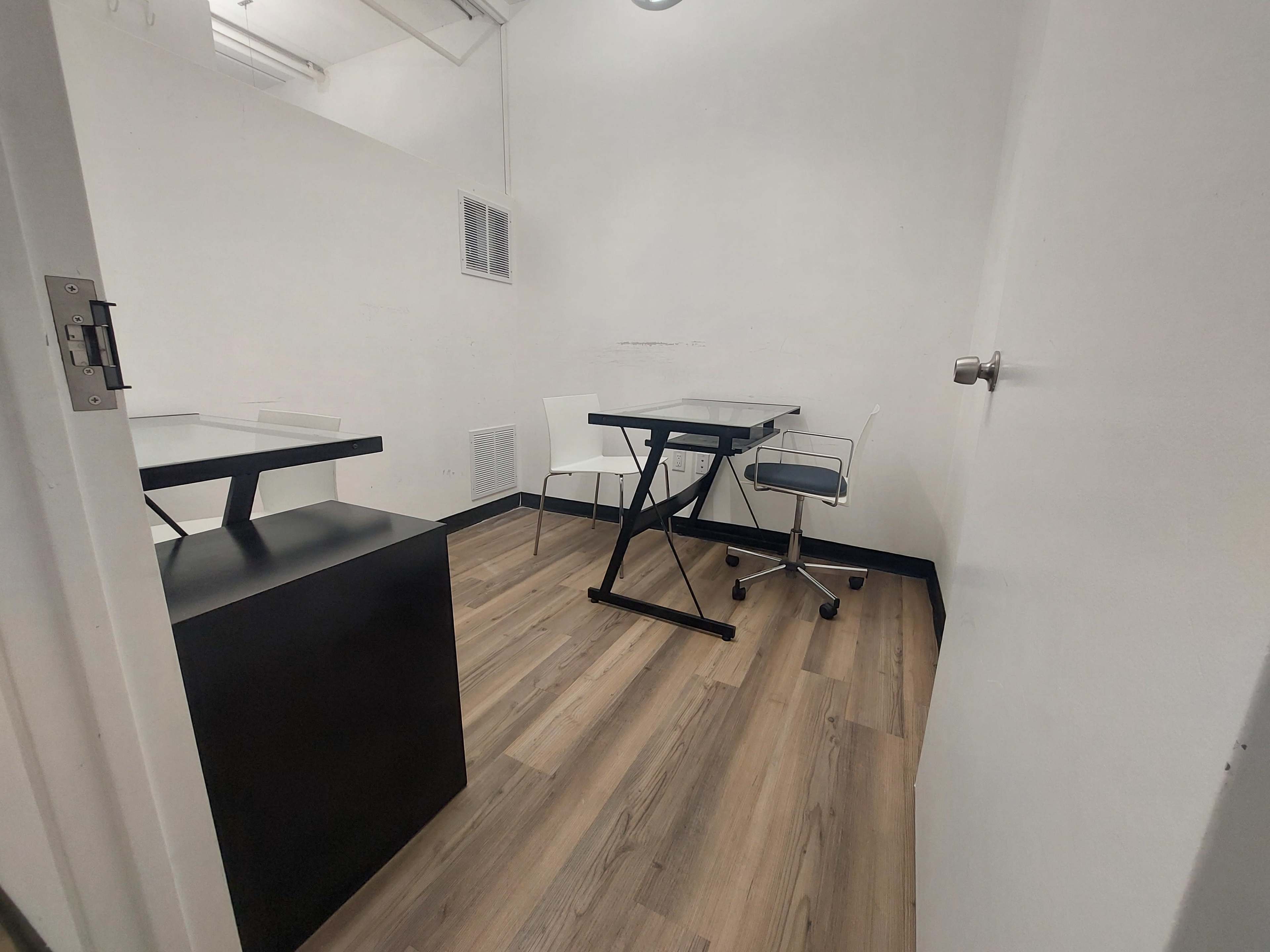 The image depicts a small, empty office space with a black desk and a chair, alongside a white table and chair, set on wooden flooring.