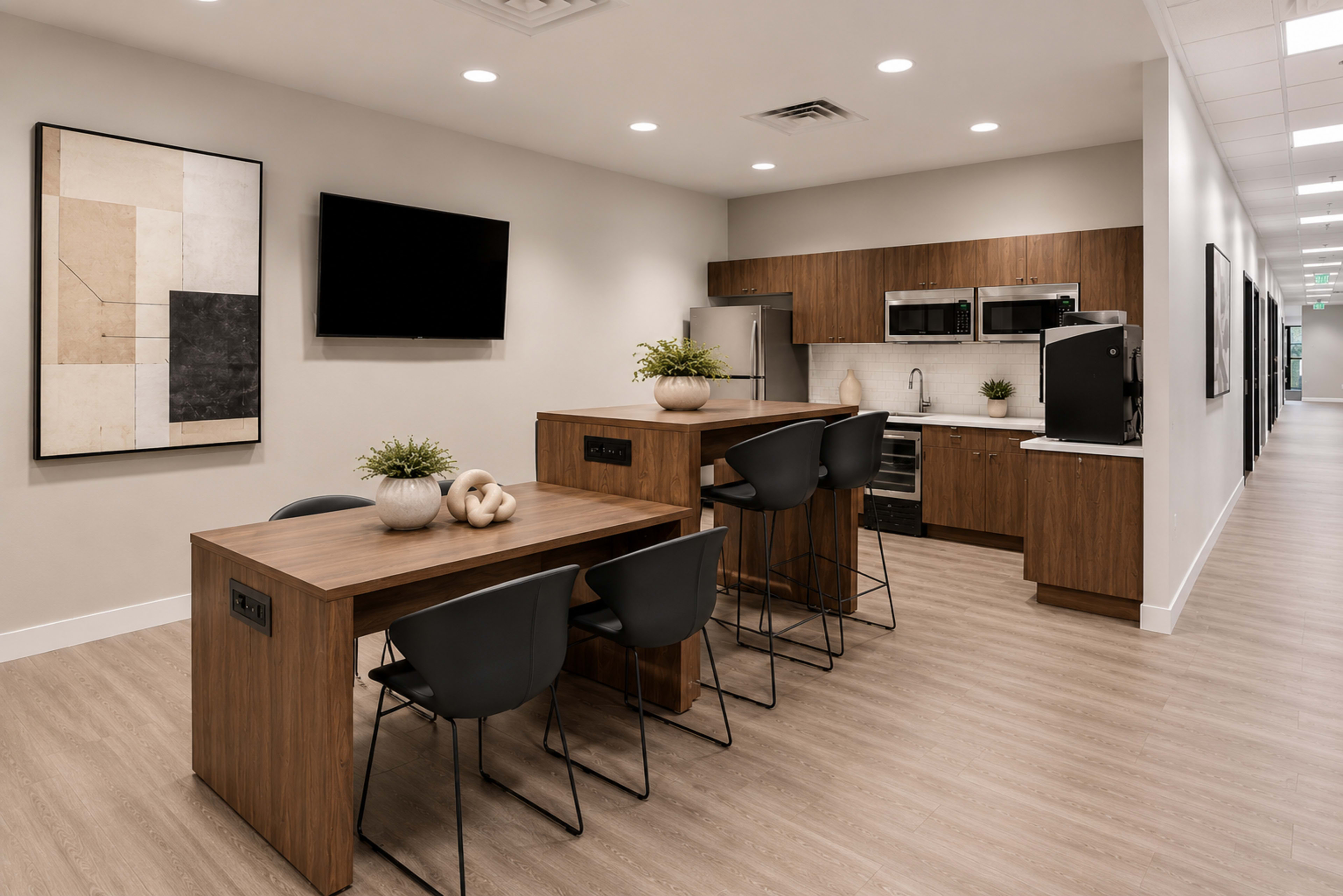 The image shows a modern kitchenette area with two wooden tables and black chairs, featuring a television on the wall and appliances such as a refrigerator and microwave.