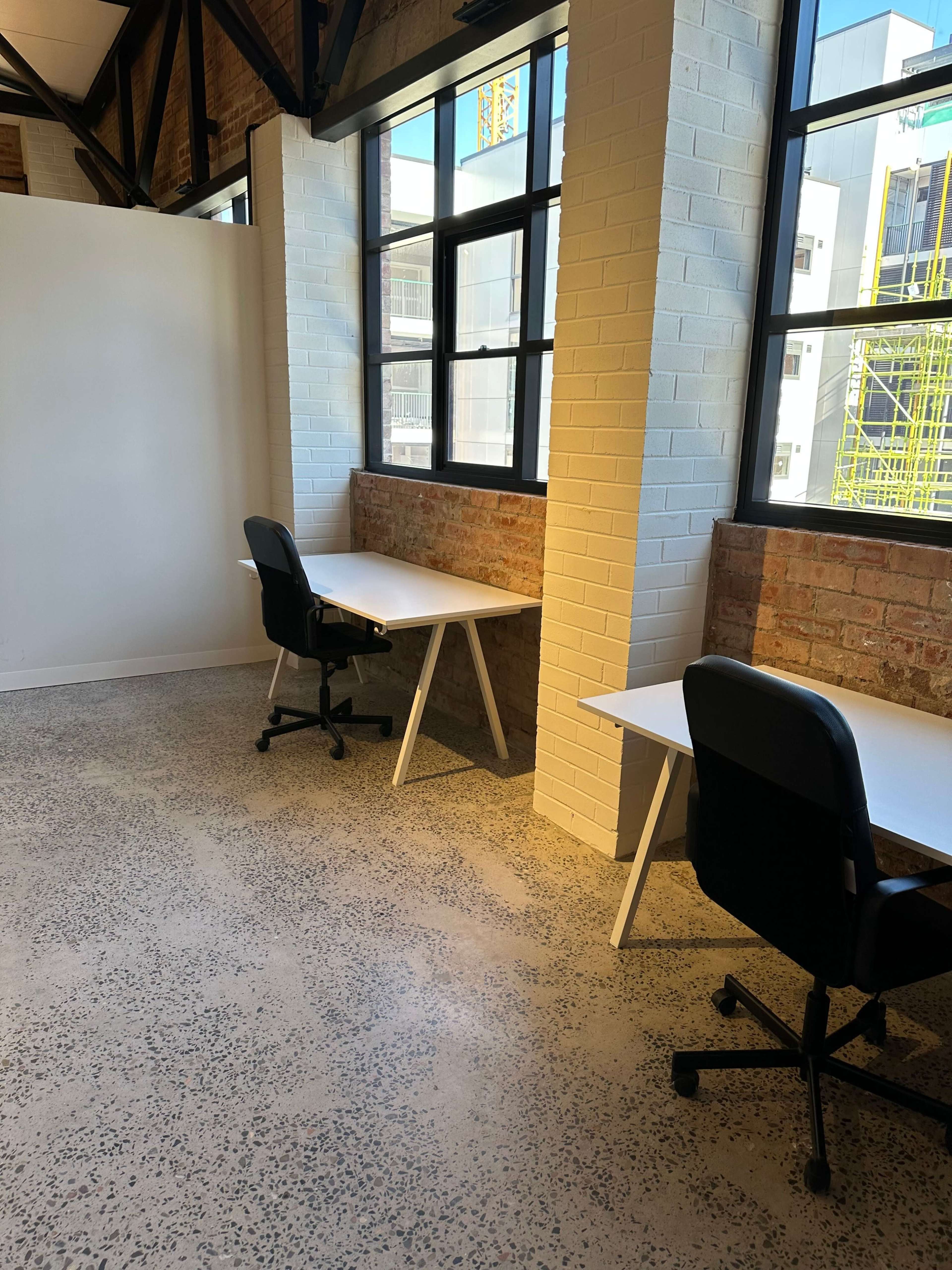 A light-filled office space featuring two desks with black chairs situated near large windows, against a backdrop of exposed brick walls.