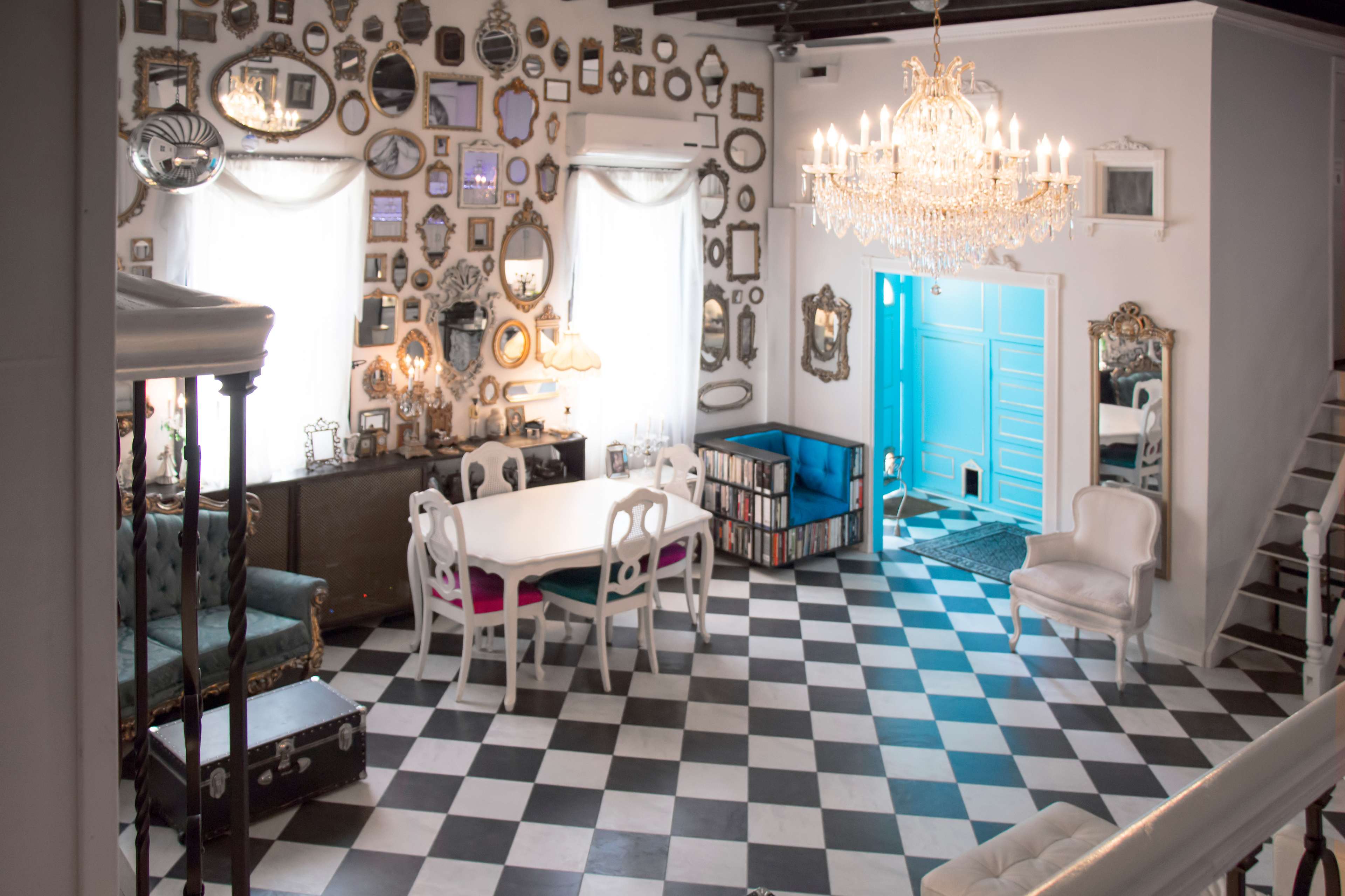A spacious room with a checkered floor, adorned with numerous decorative mirrors on the walls, a large chandelier overhead, and a bright blue door leading outside.