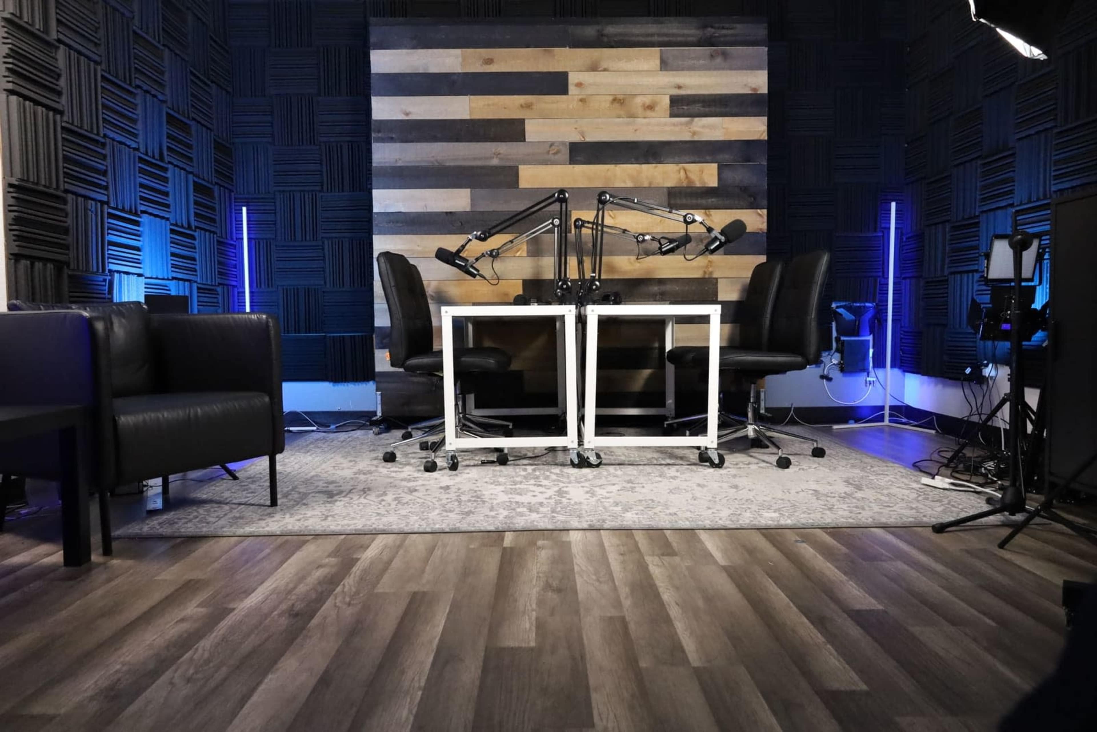 A modern podcast studio with soundproofed walls, two microphone stands positioned over a table, and dark seating arrangements.