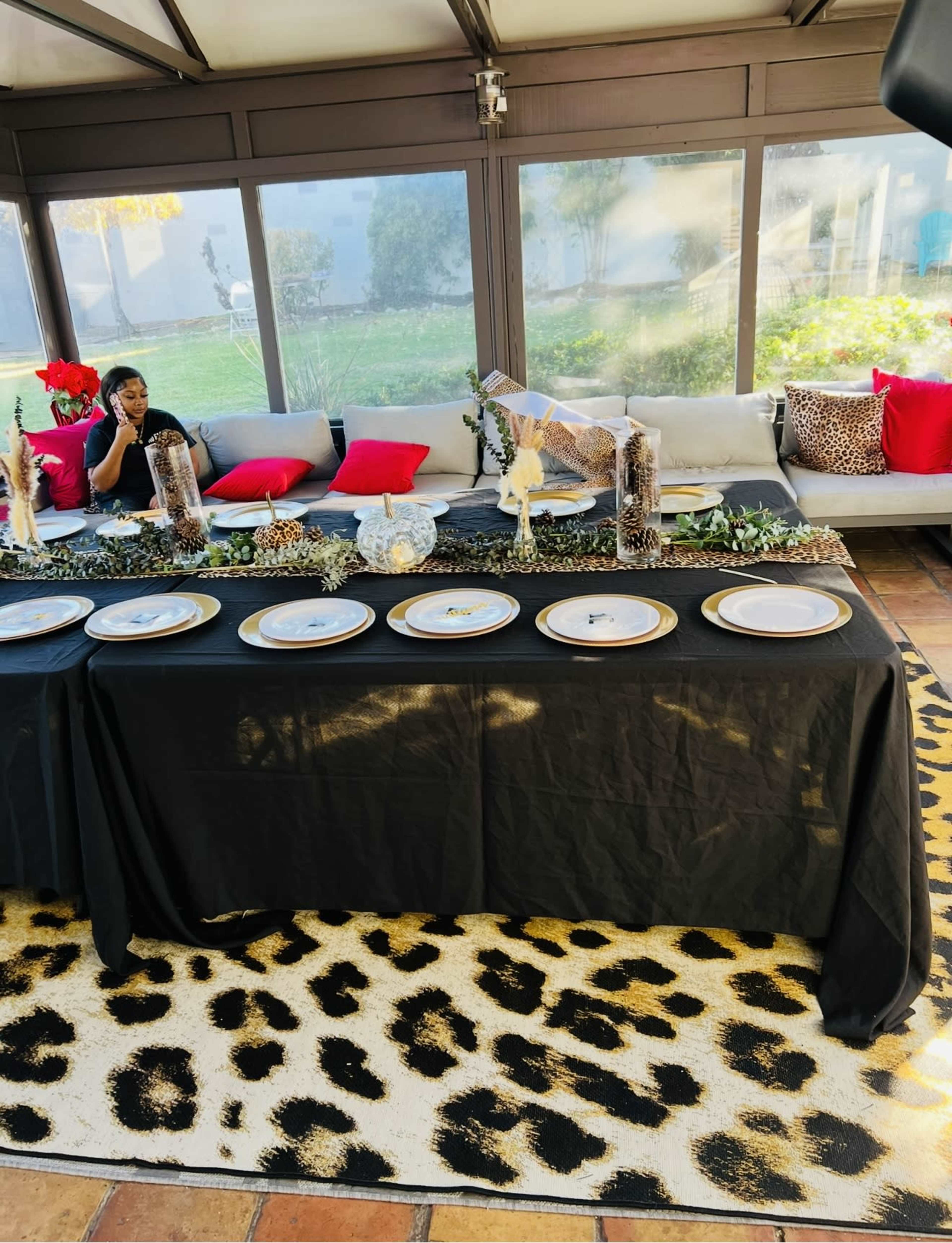 A table set with several plates is arranged in a sunroom decorated with a leopard-print rug, while a person sits on a couch adorned with colorful pillows in the background.