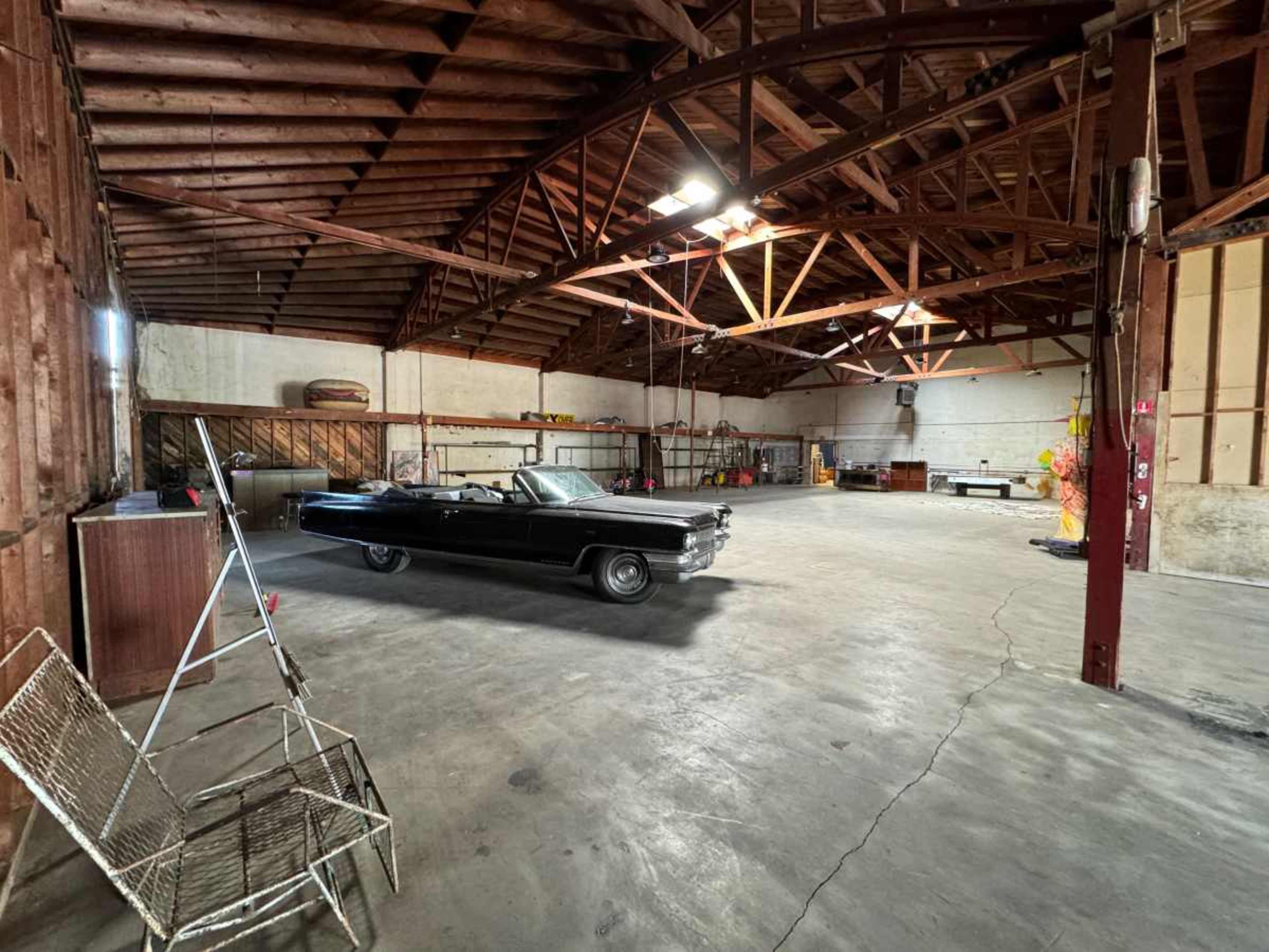 The image shows a spacious, empty warehouse with a vintage black car parked in the center and a metal folding chair in the foreground.