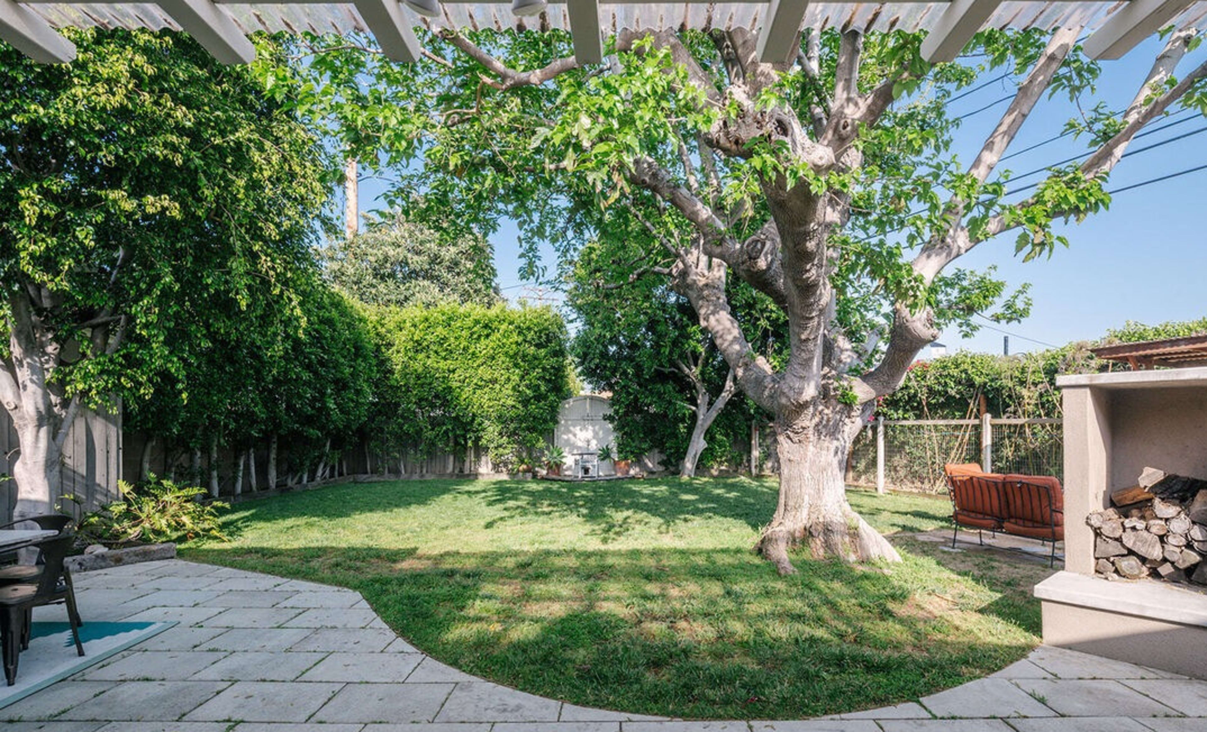 A spacious backyard features a large tree and manicured grass surrounded by tall hedges and a stone patio.