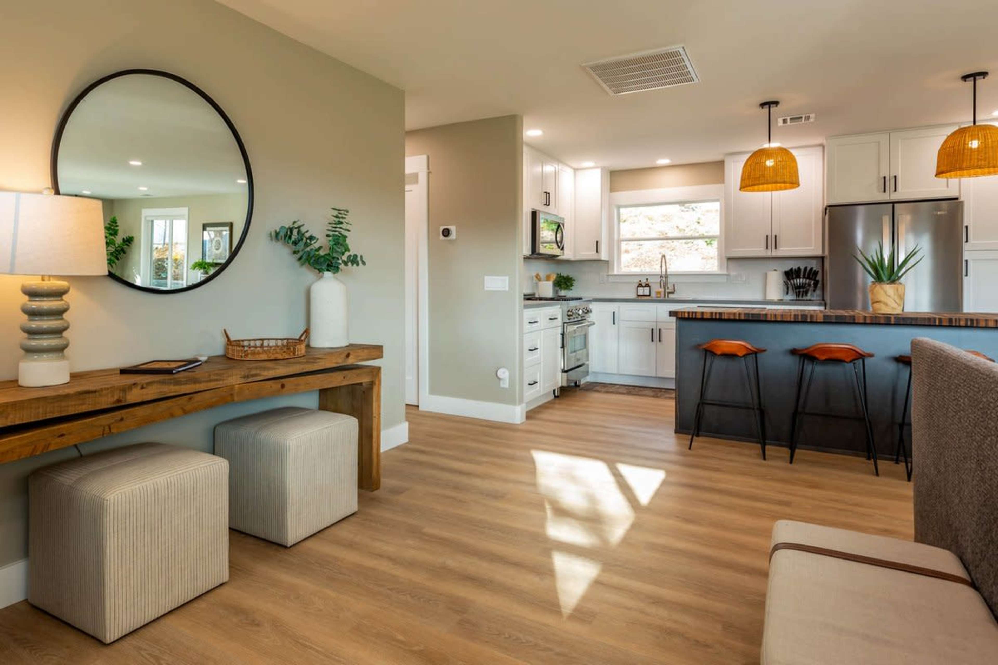 A modern kitchen and living area featuring a wooden table with two stools, a large circular mirror, and pendant lighting above a kitchen island.