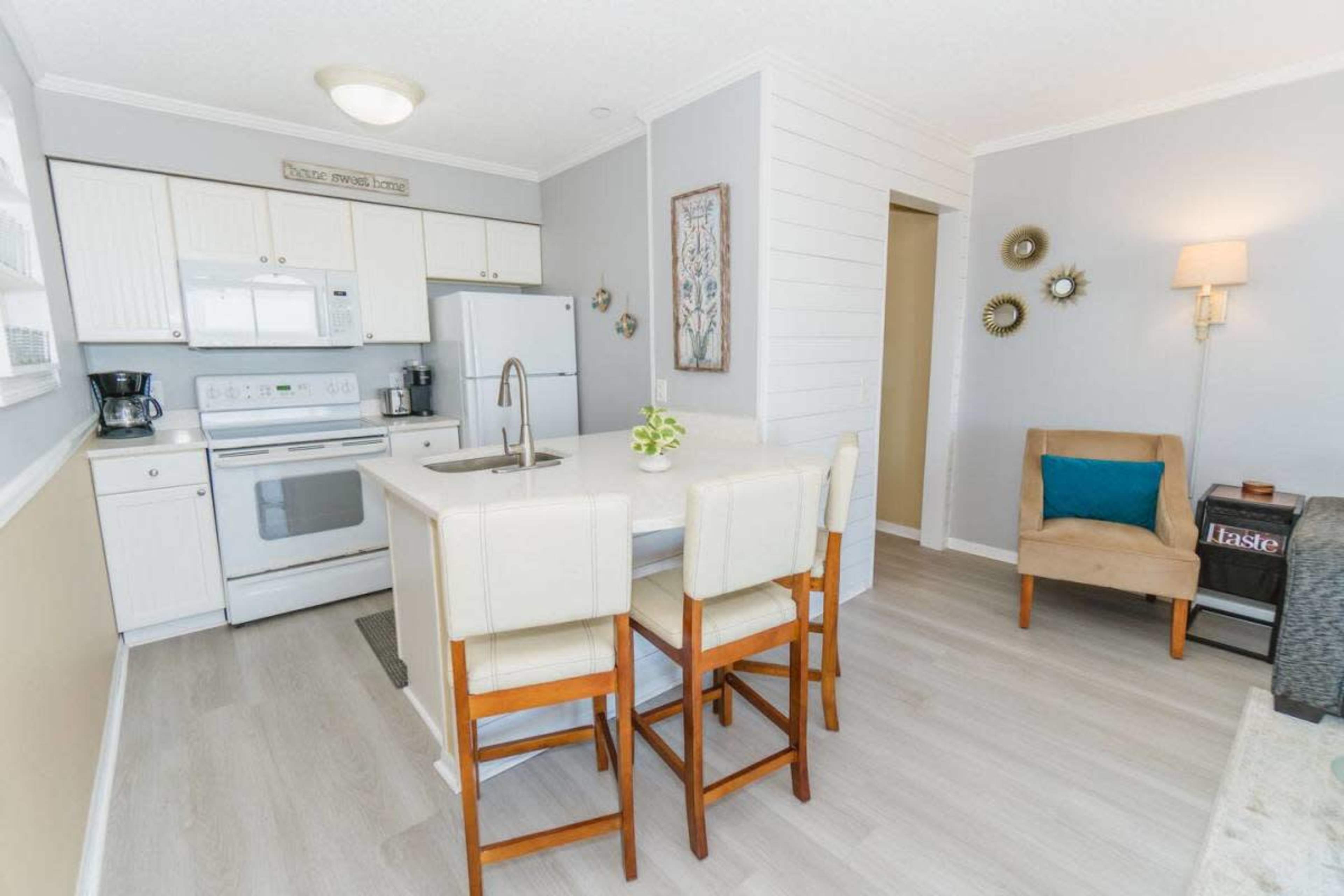 The image shows a modern kitchen with white cabinetry, a small island with bar stools, and a cozy seating area to the right.