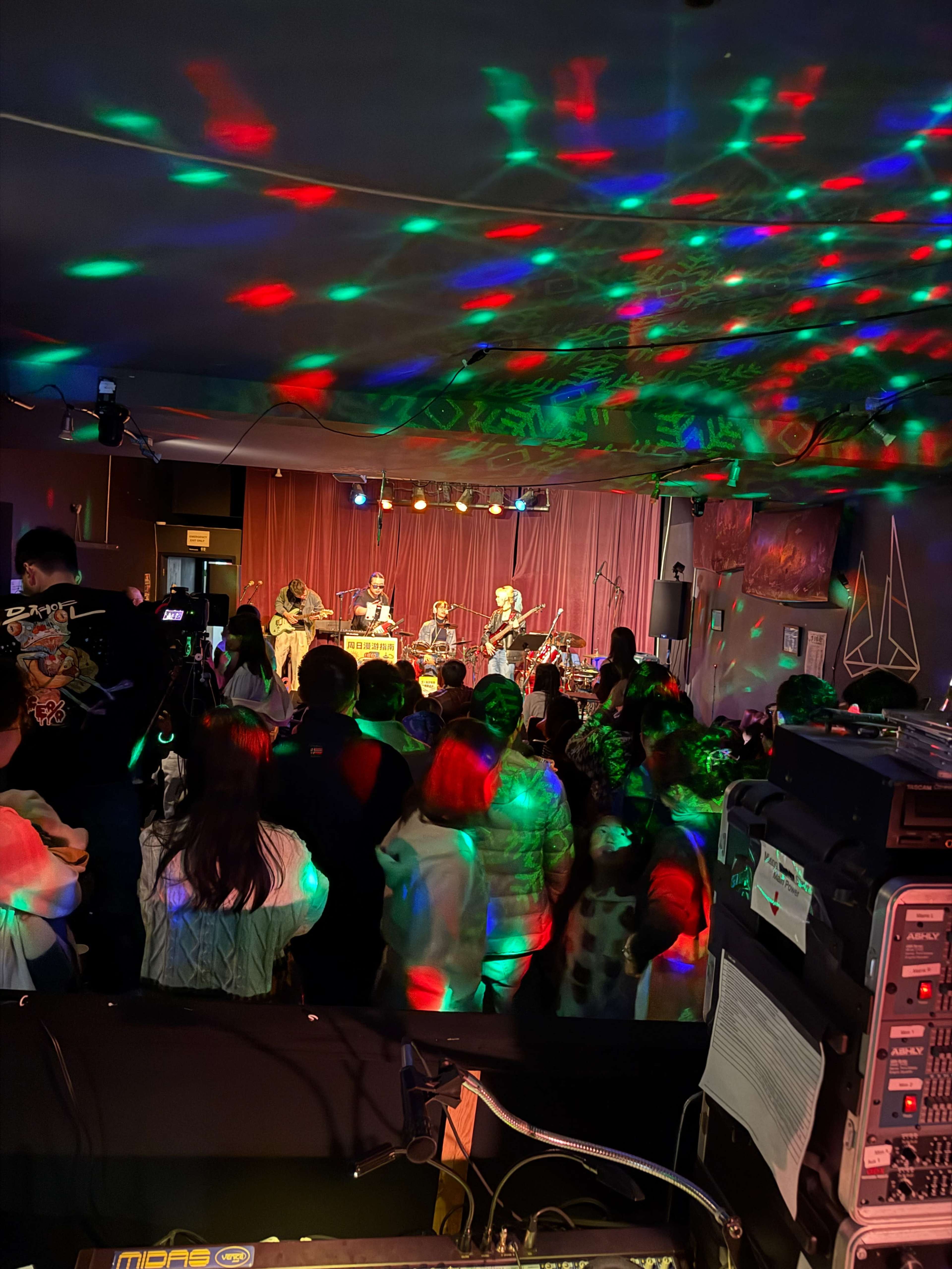 A band performs on stage while colorful lights illuminate a crowd of people dancing and enjoying the show.