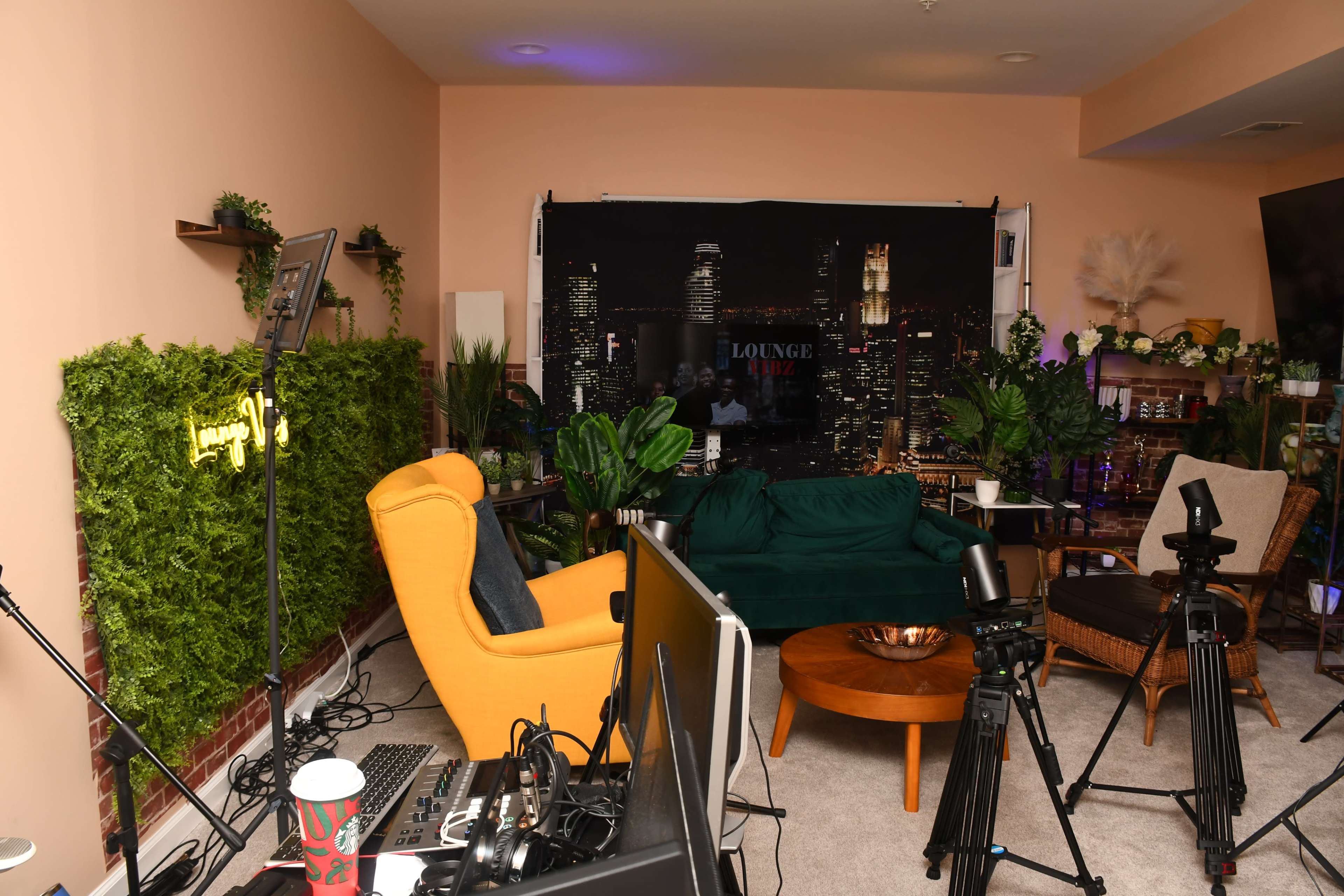 The image shows a cozy lounge area with green plants, two sofas, a round coffee table, and filming equipment set up in a well-lit room.