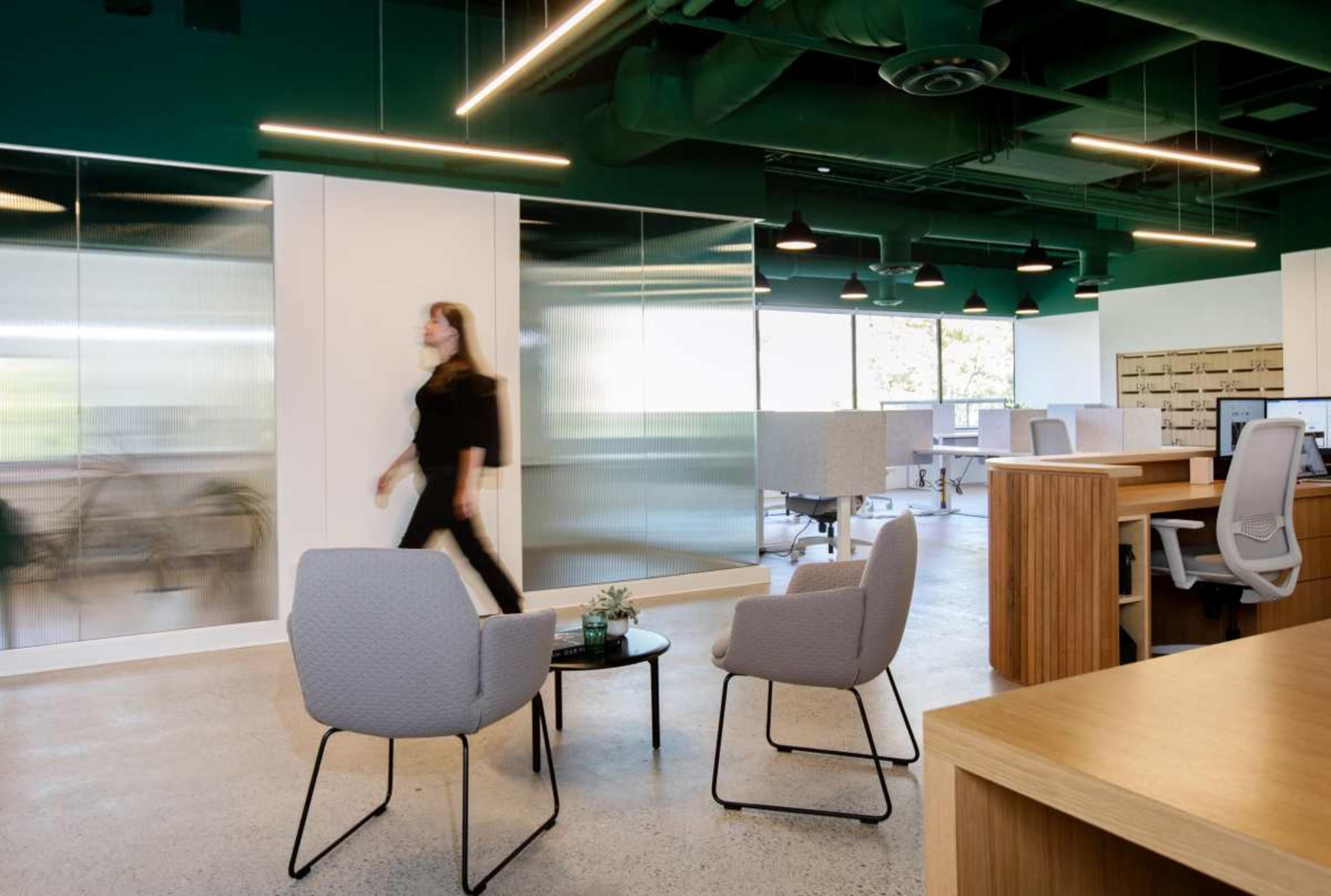 A woman walks through a modern office space featuring gray chairs, a coffee table, and glass partition walls.