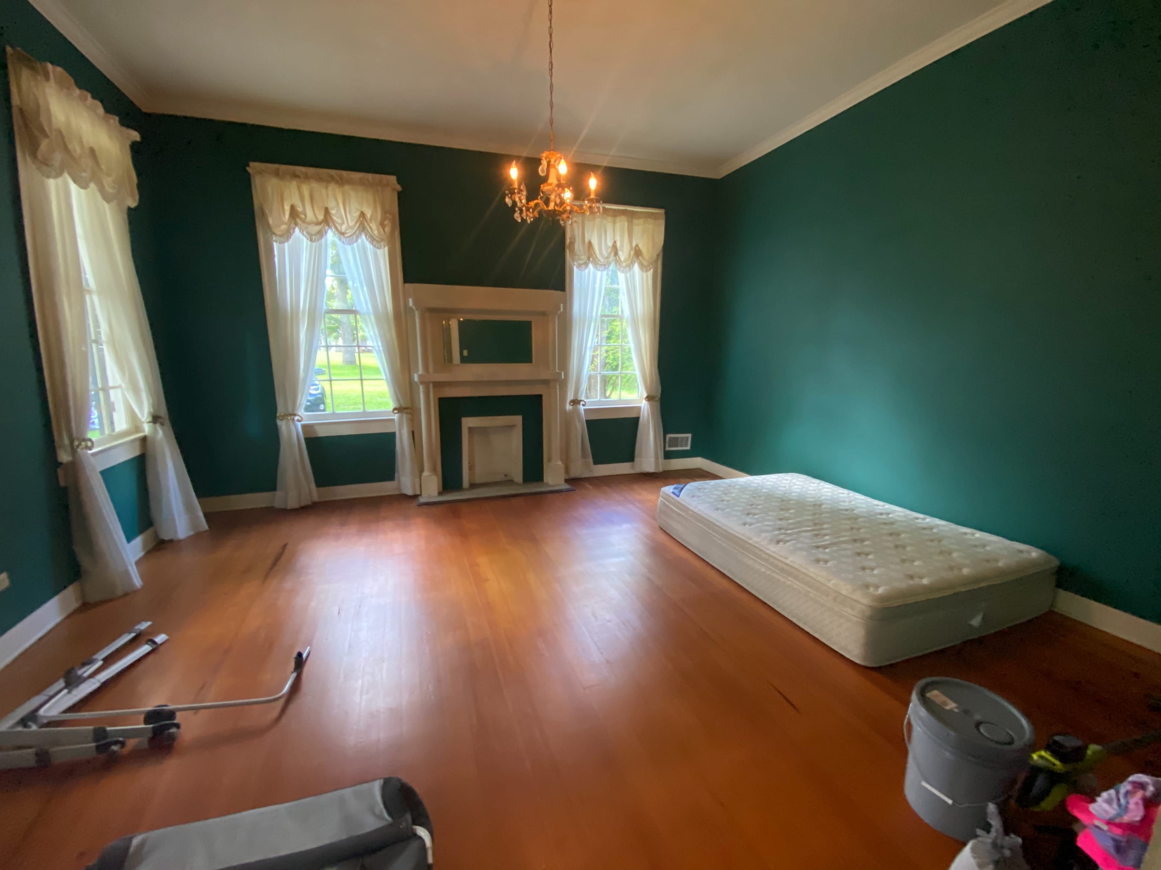 The room features green walls, a central chandelier, a fireplace mantel, two large windows with drapes, a mattress on the wooden floor, and some scattered items in the corner.
