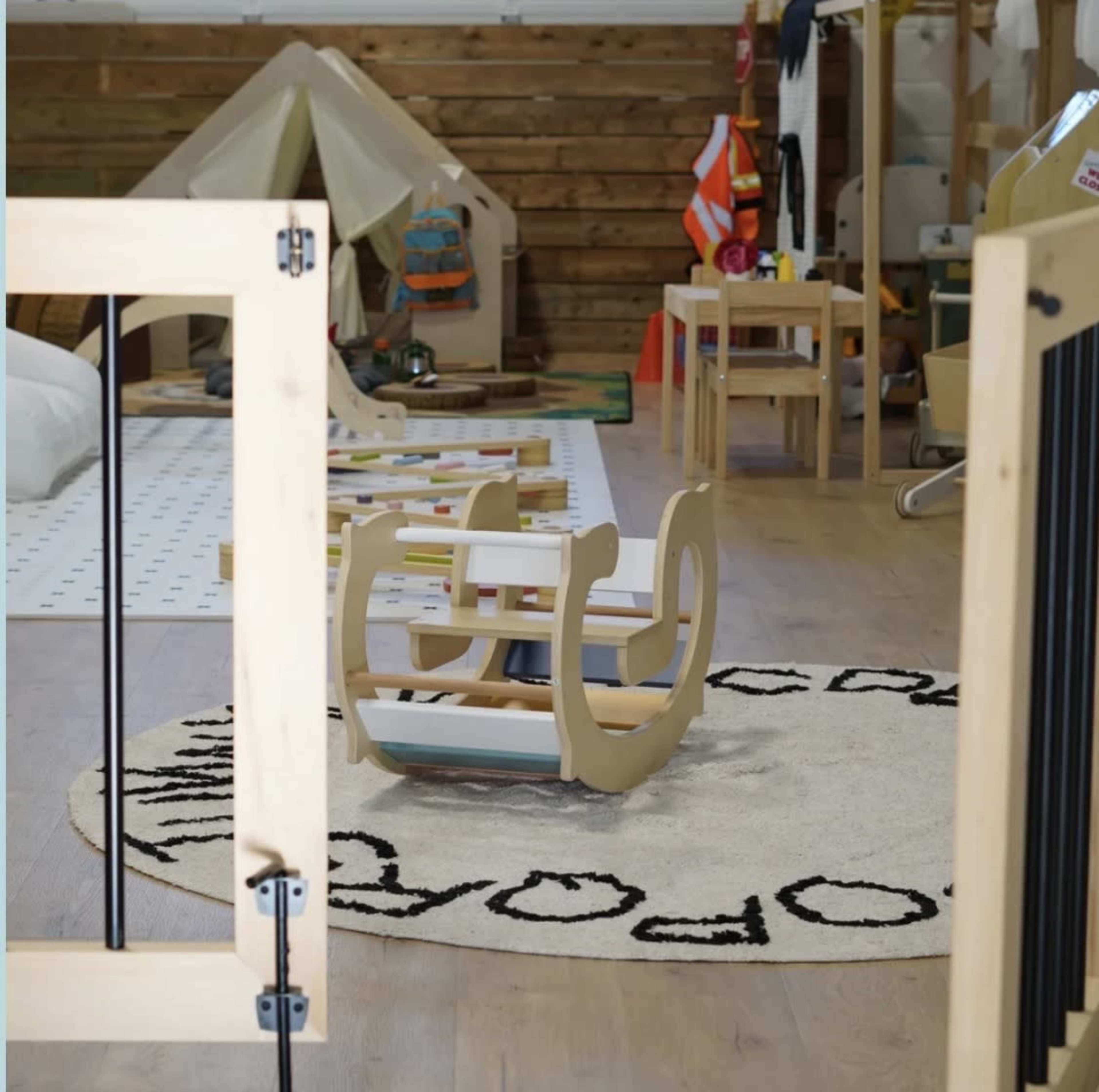 The image shows a play area featuring a colorful rocking chair on a round rug, with wooden furniture and a tent in the background.