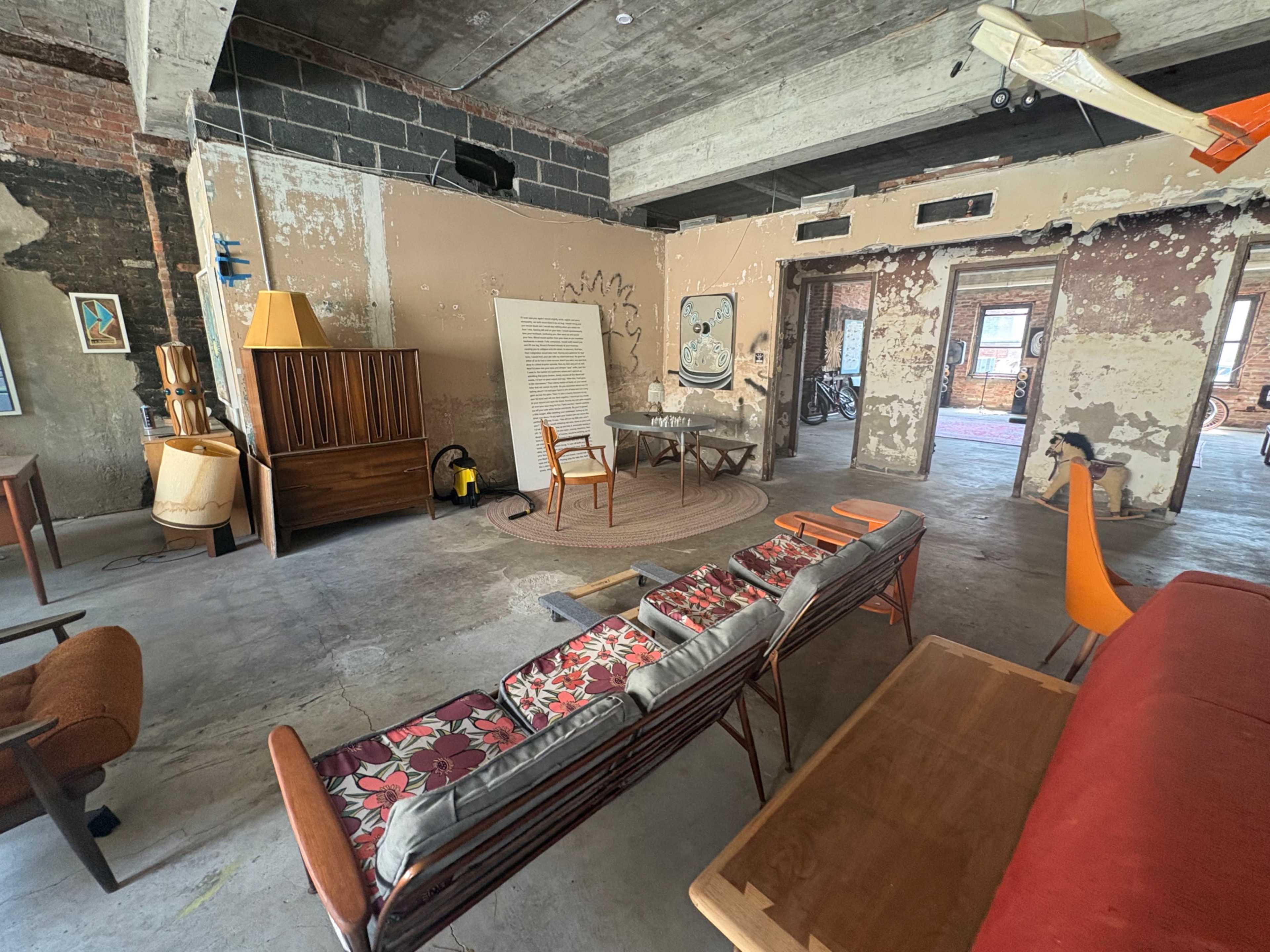 The image shows a spacious, industrial-style room with exposed brick walls, various pieces of retro furniture, and a large window allowing natural light to enter.