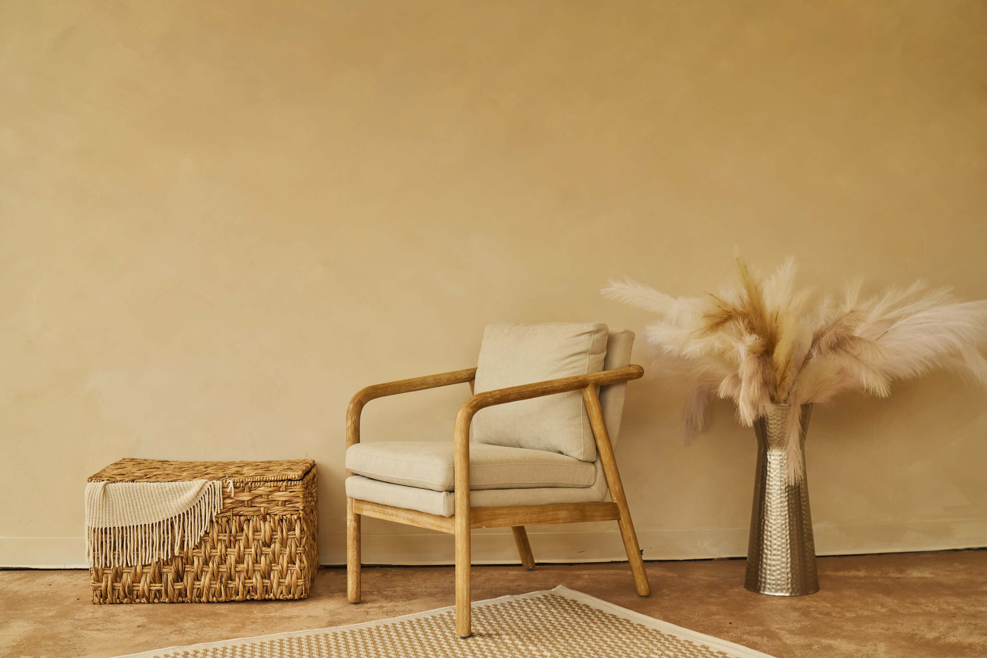 A wooden chair with a light cushion is positioned next to a woven basket and a vase filled with dried pampas grass against a beige wall.