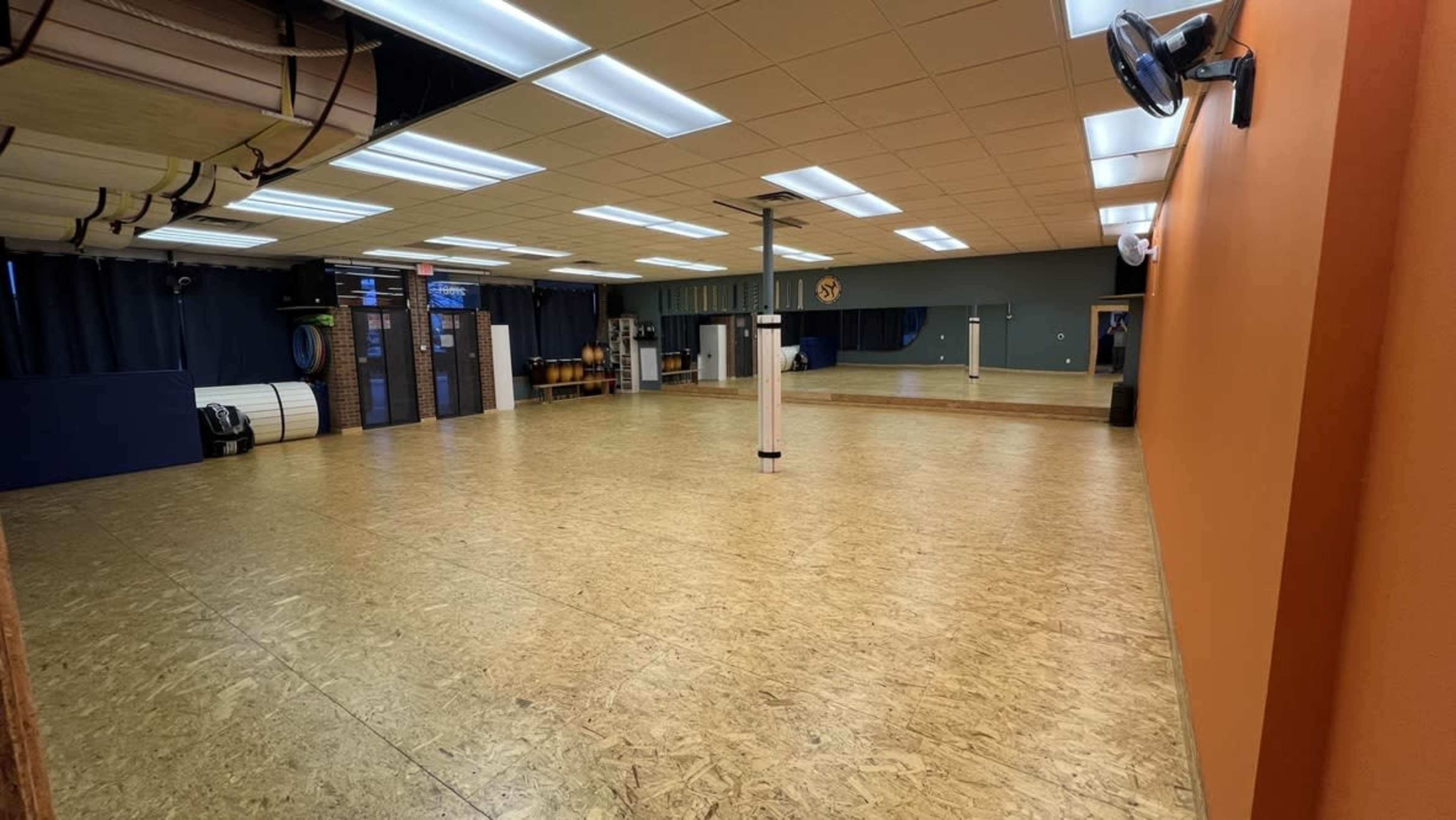 A spacious dance studio with wooden flooring, mirrors on one wall, and various equipment neatly arranged along the sides.