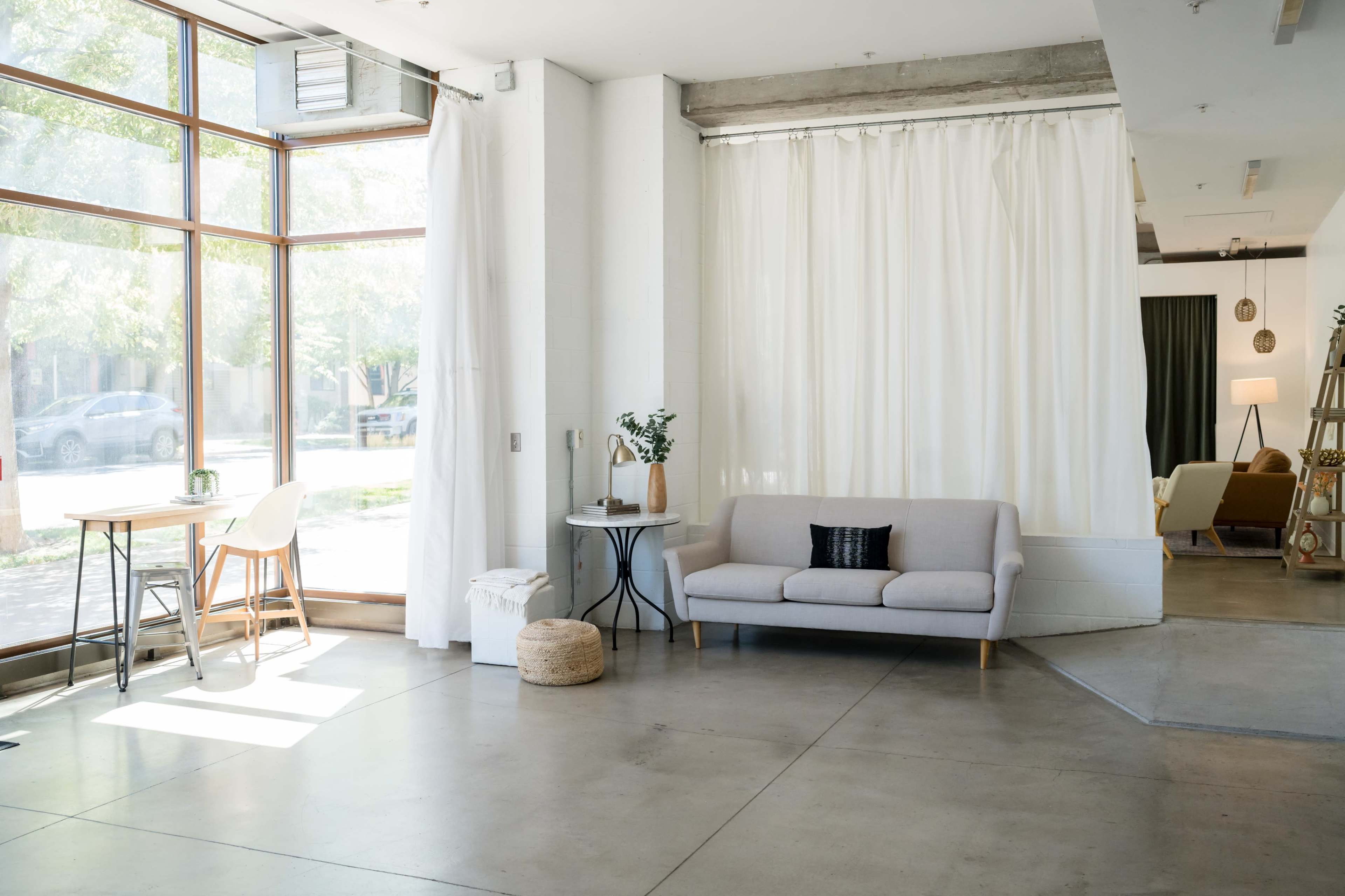 The interior features a bright space with large windows, a sofa, a small table with chairs, and white curtains draping across one section.