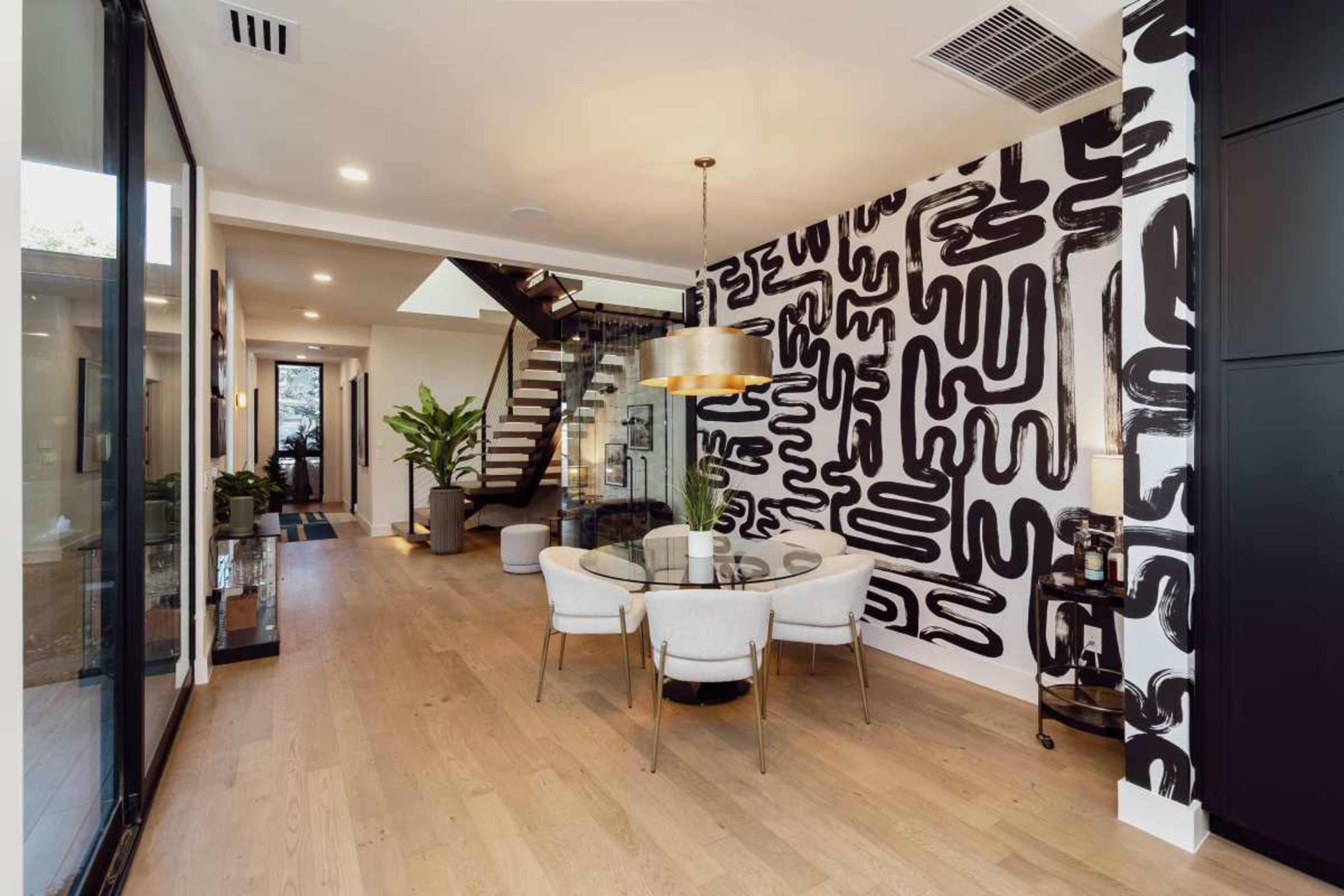 The image shows a modern entryway with a staircase, a round dining table surrounded by four chairs, and a large abstract black and white mural on one wall.