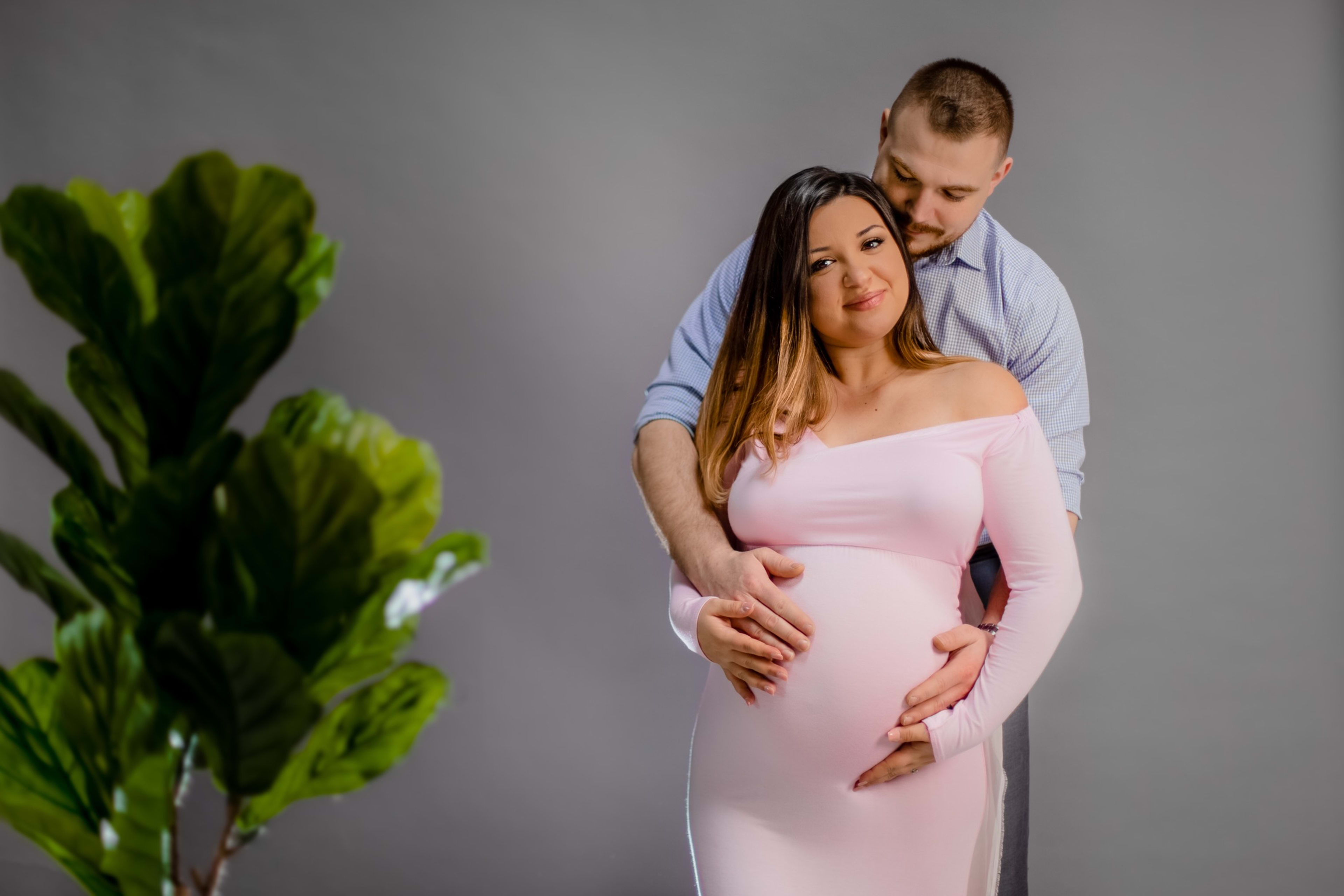 A pregnant woman in a pink dress poses for a maternity photo shoot with her husband.