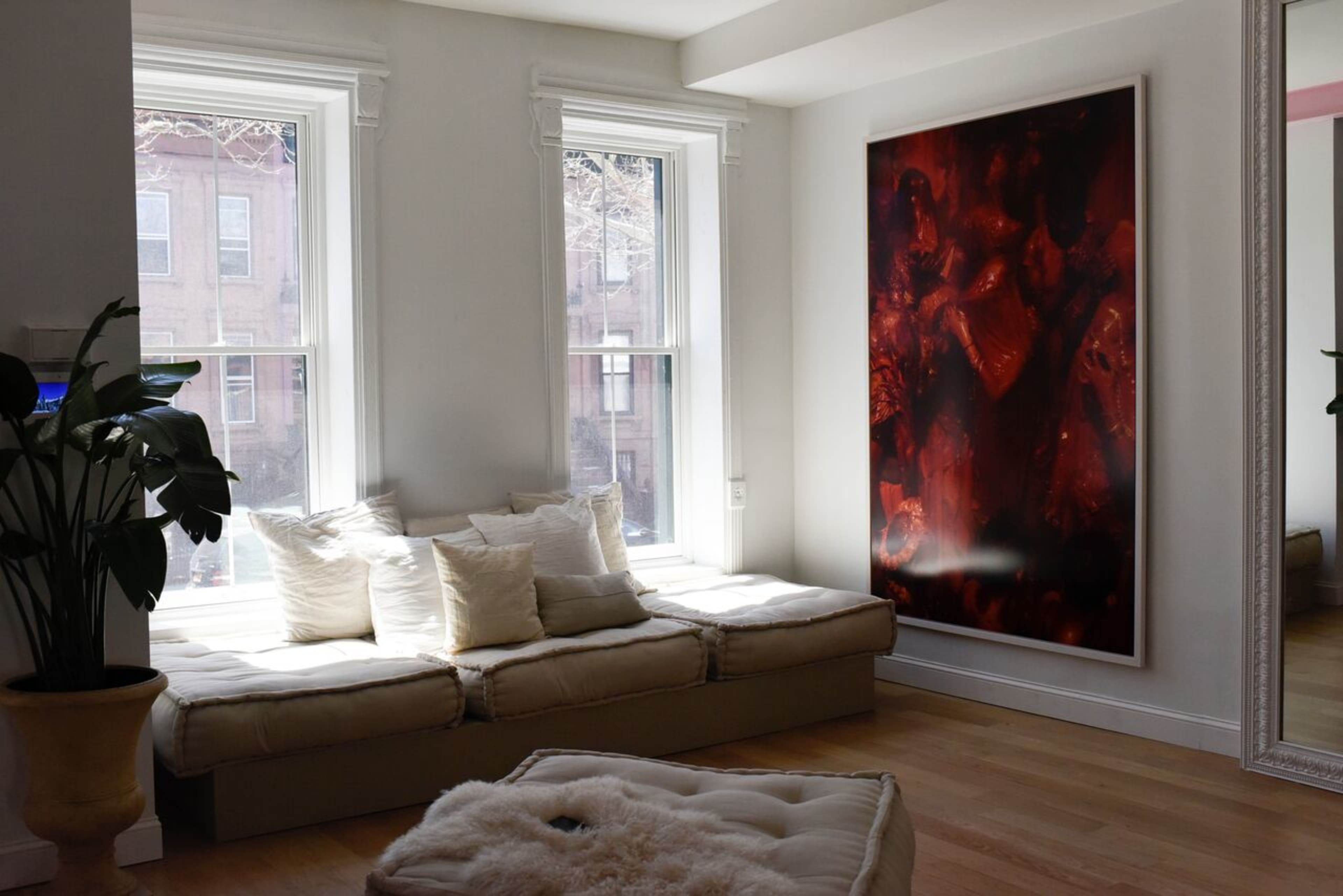 A bright room with two large windows, a cushioned seating area against the wall, and a large artwork hanging beside a mirror.