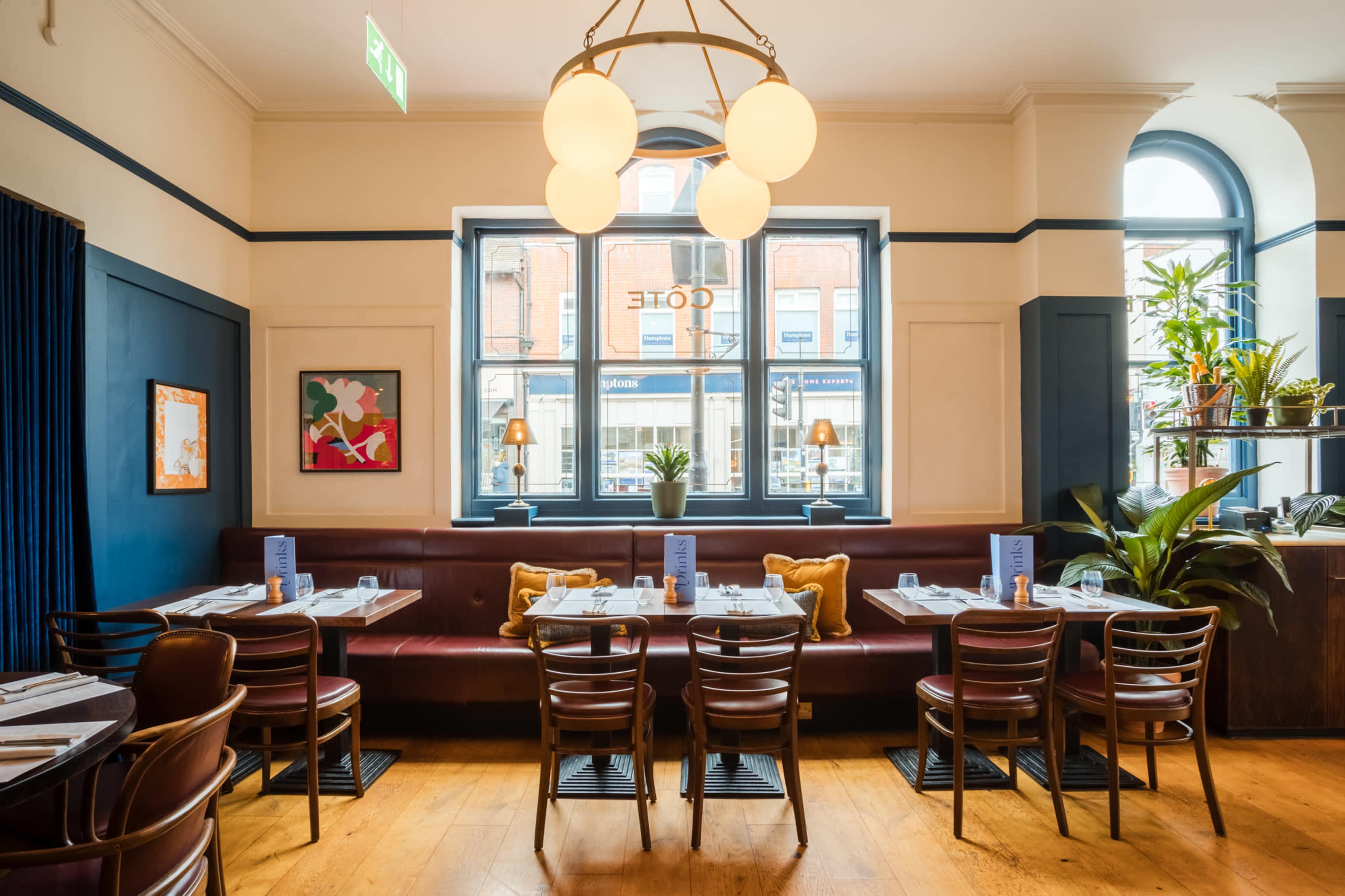The image shows a bright restaurant interior with wooden flooring, a maroon banquette, and several tables set with menus and dining utensils.
