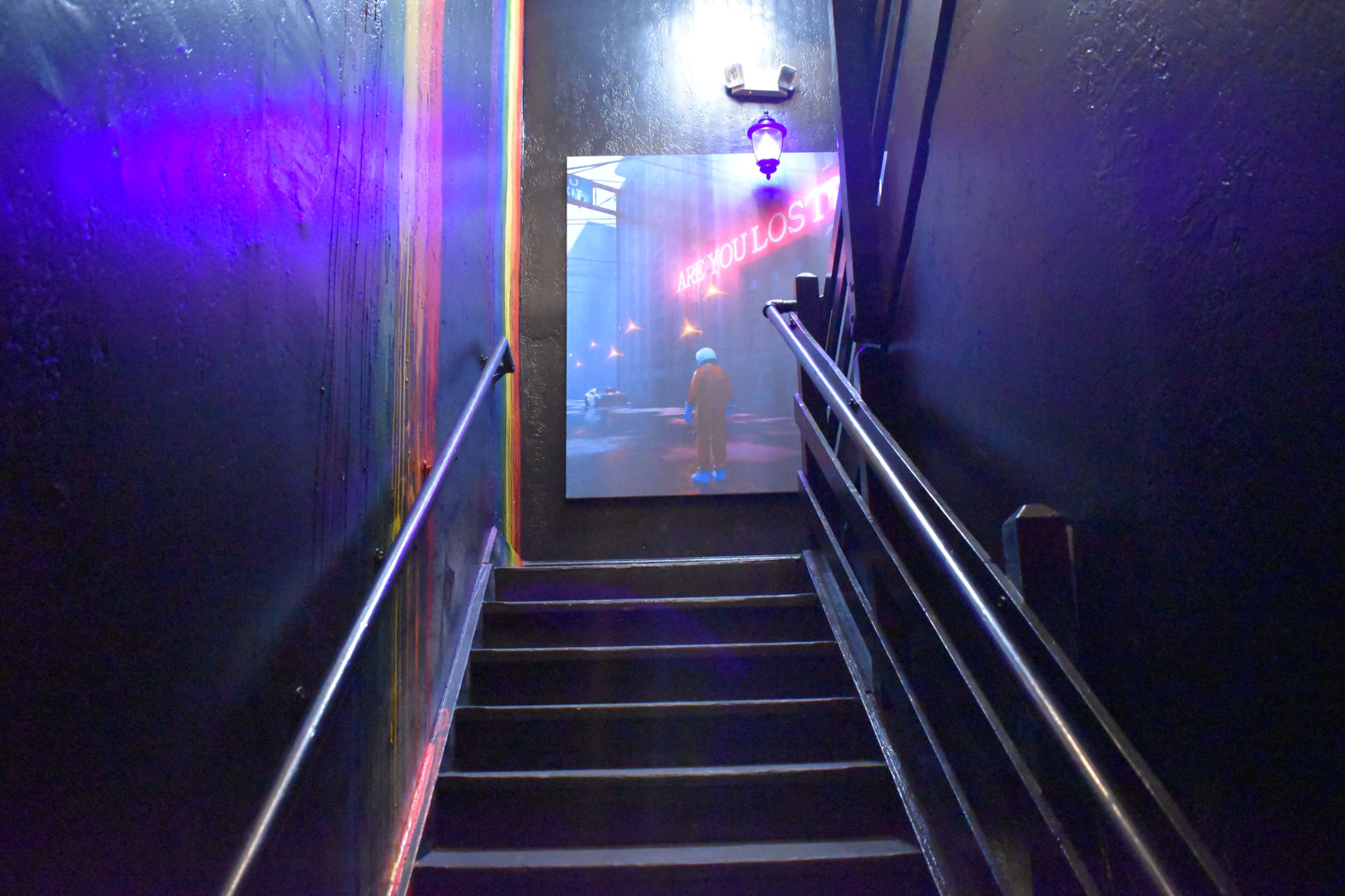 A dimly lit staircase leads down to a large framed image on the wall that features the words "ARE YOU LOST?" illuminated in neon lights.