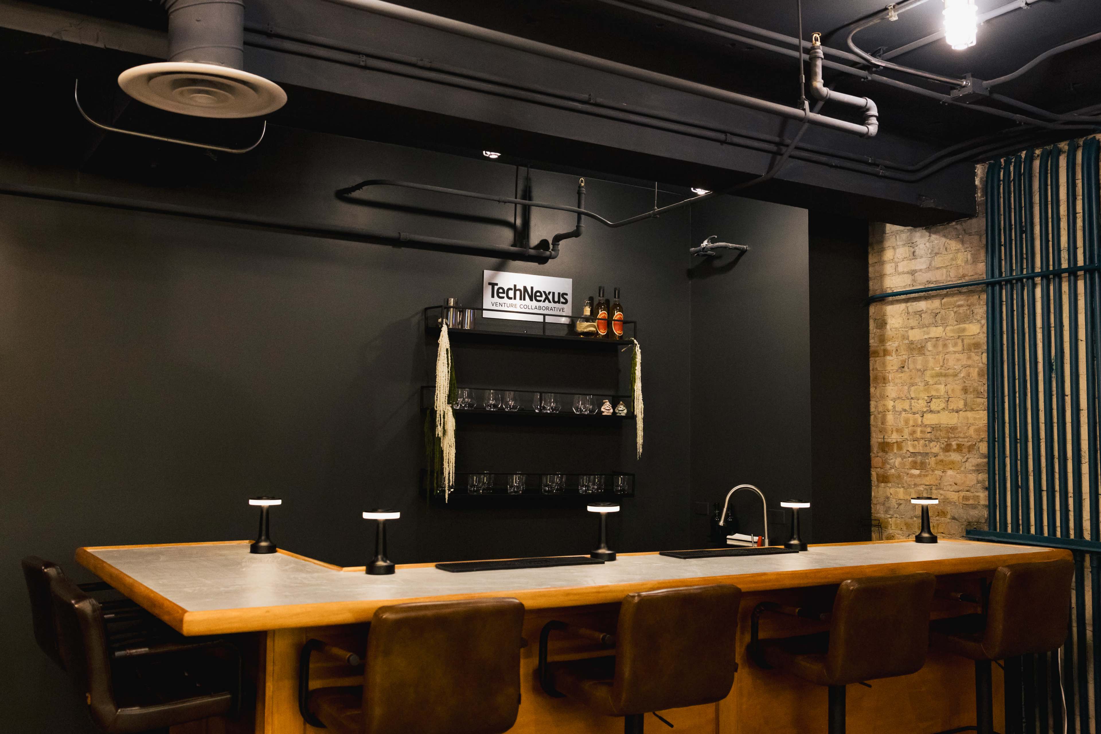The image shows a modern bar area with a wooden counter, several barstools, and a shelf displaying drinks against a dark wall.