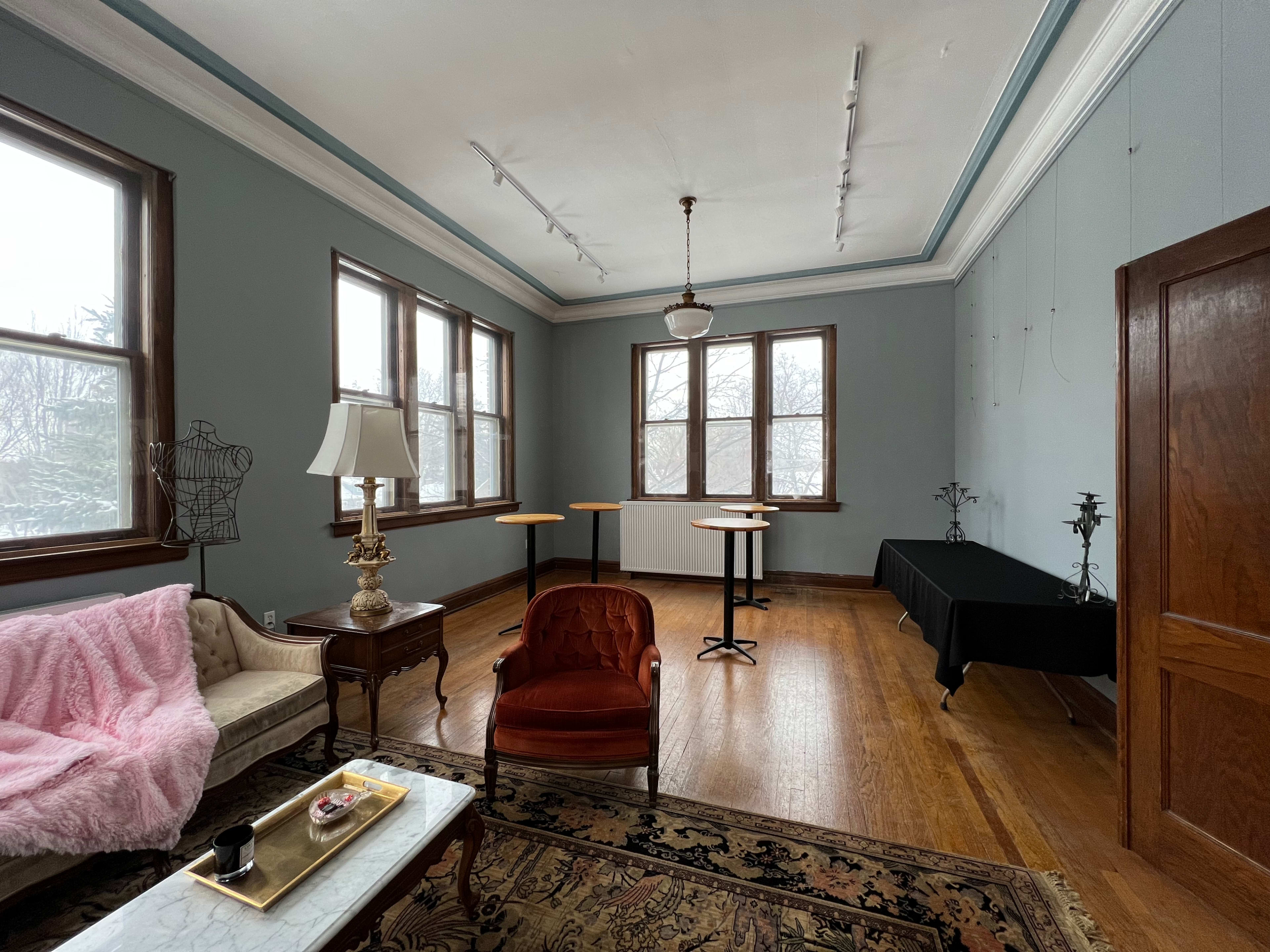The room features large windows with a view of a gray day, a beige sofa with a pink throw, a red armchair, and several small tables arranged throughout the space.