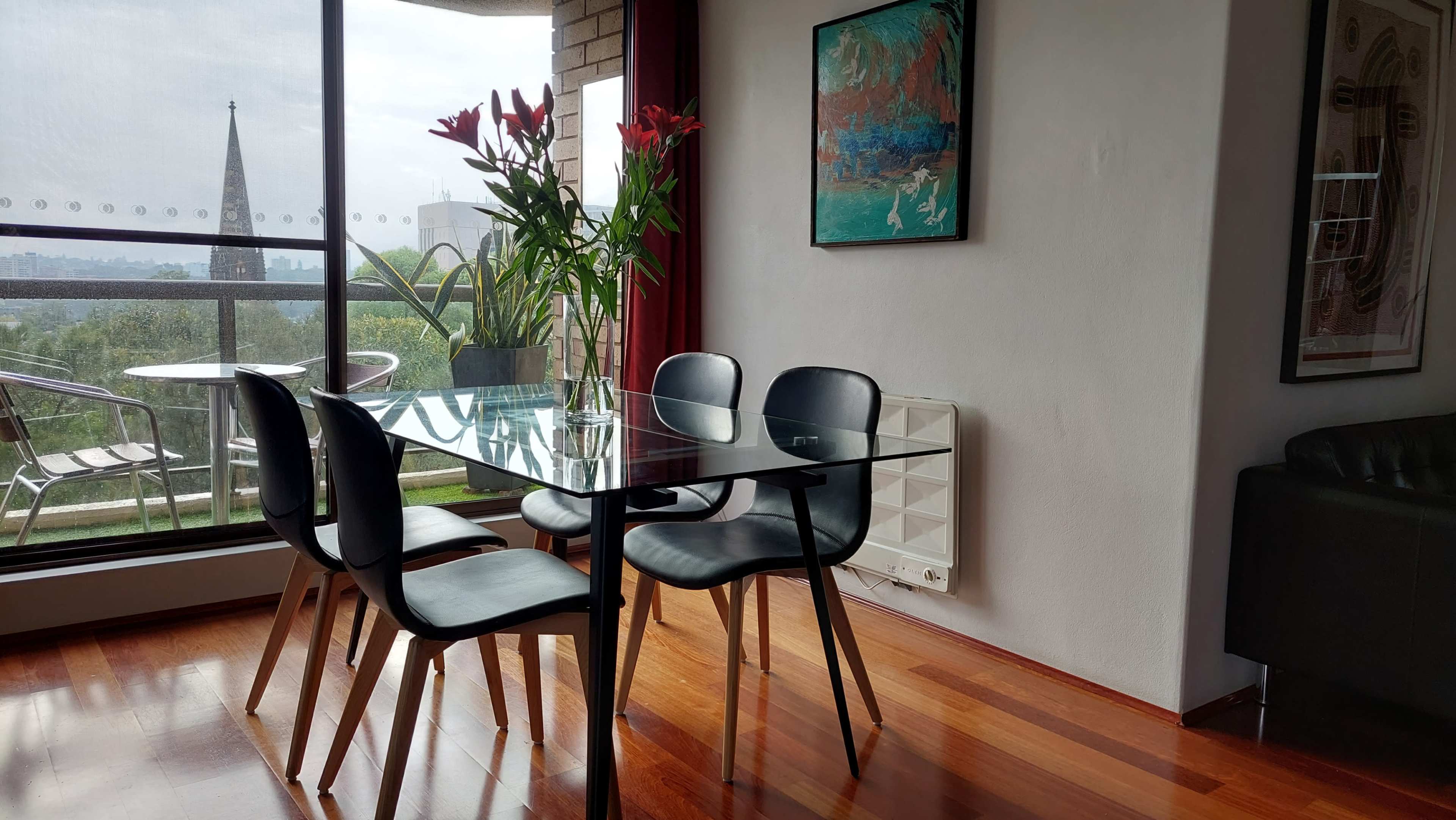 A glass dining table with four black chairs is positioned in a room with a large window overlooking a balcony and a view of a church spire in the distance.