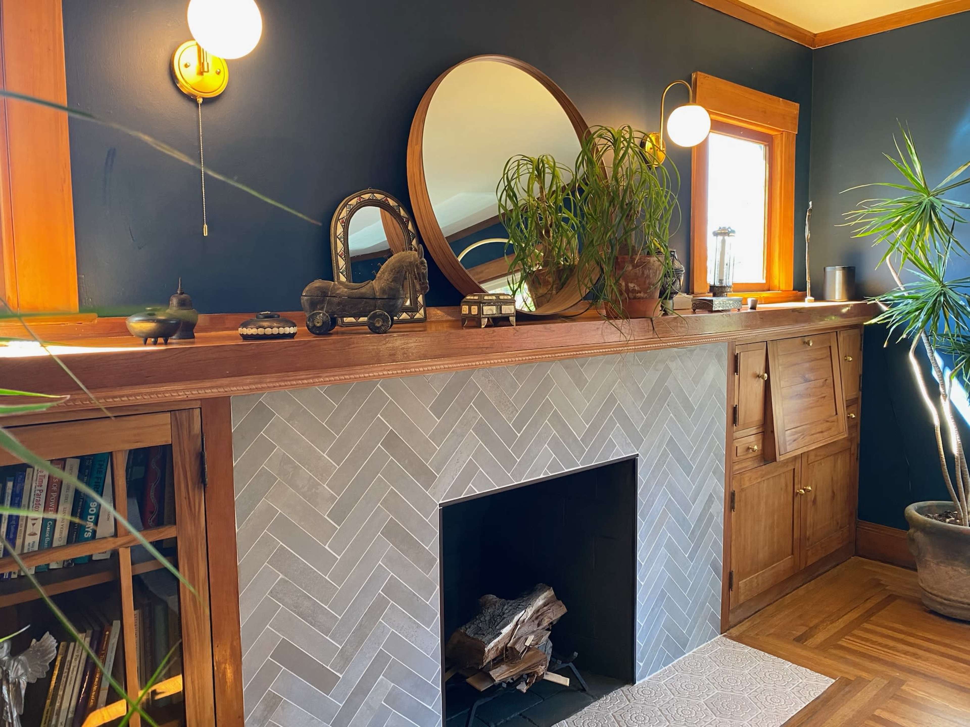 The image shows a modern living room with a herringbone-patterned fireplace, a circular mirror above it, and various decorations on a wooden mantel.