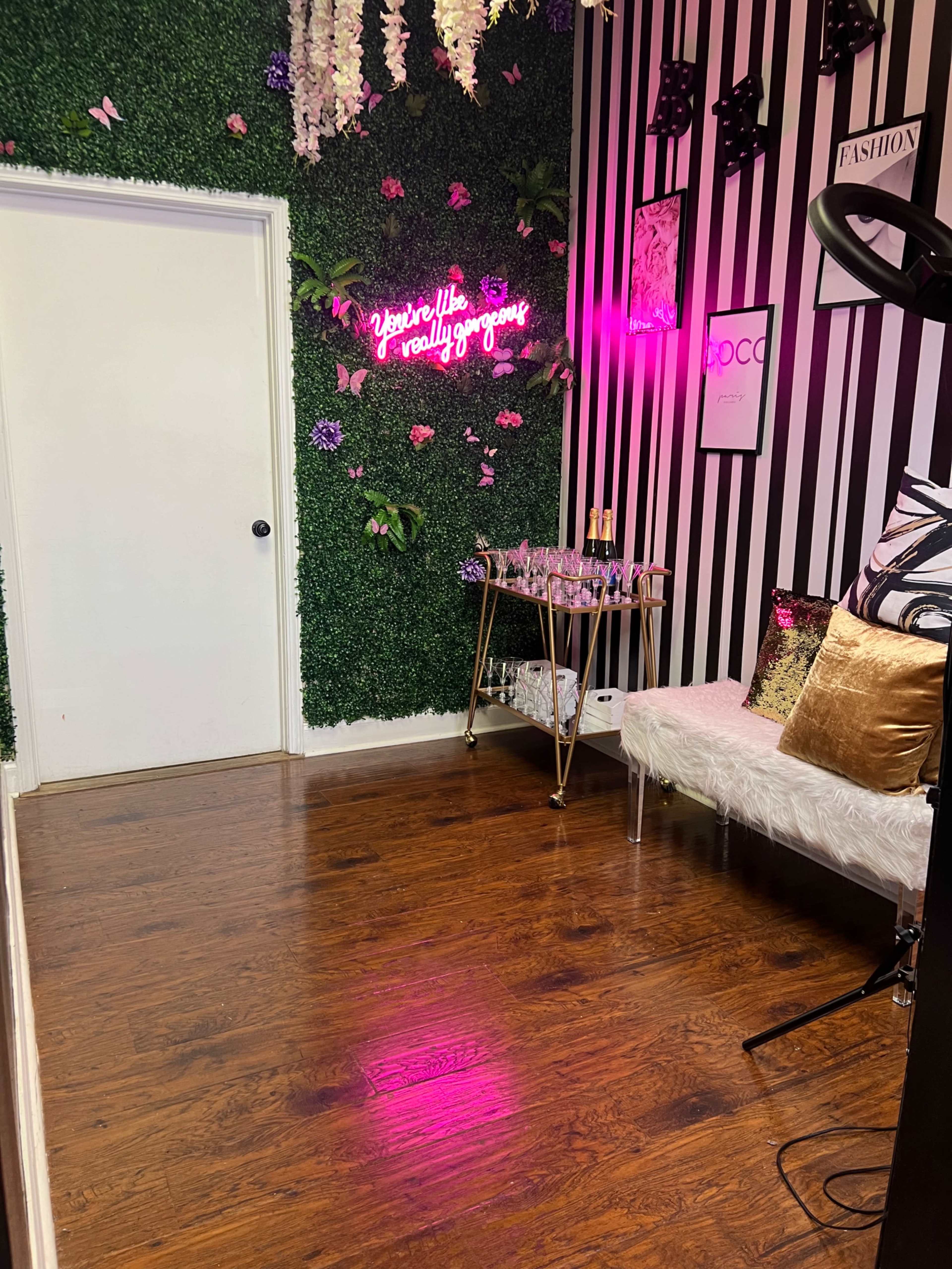 The image shows a small room with a green textured wall, a neon sign, a gold bar cart with glasses, and decorative elements like flowers and pillows.