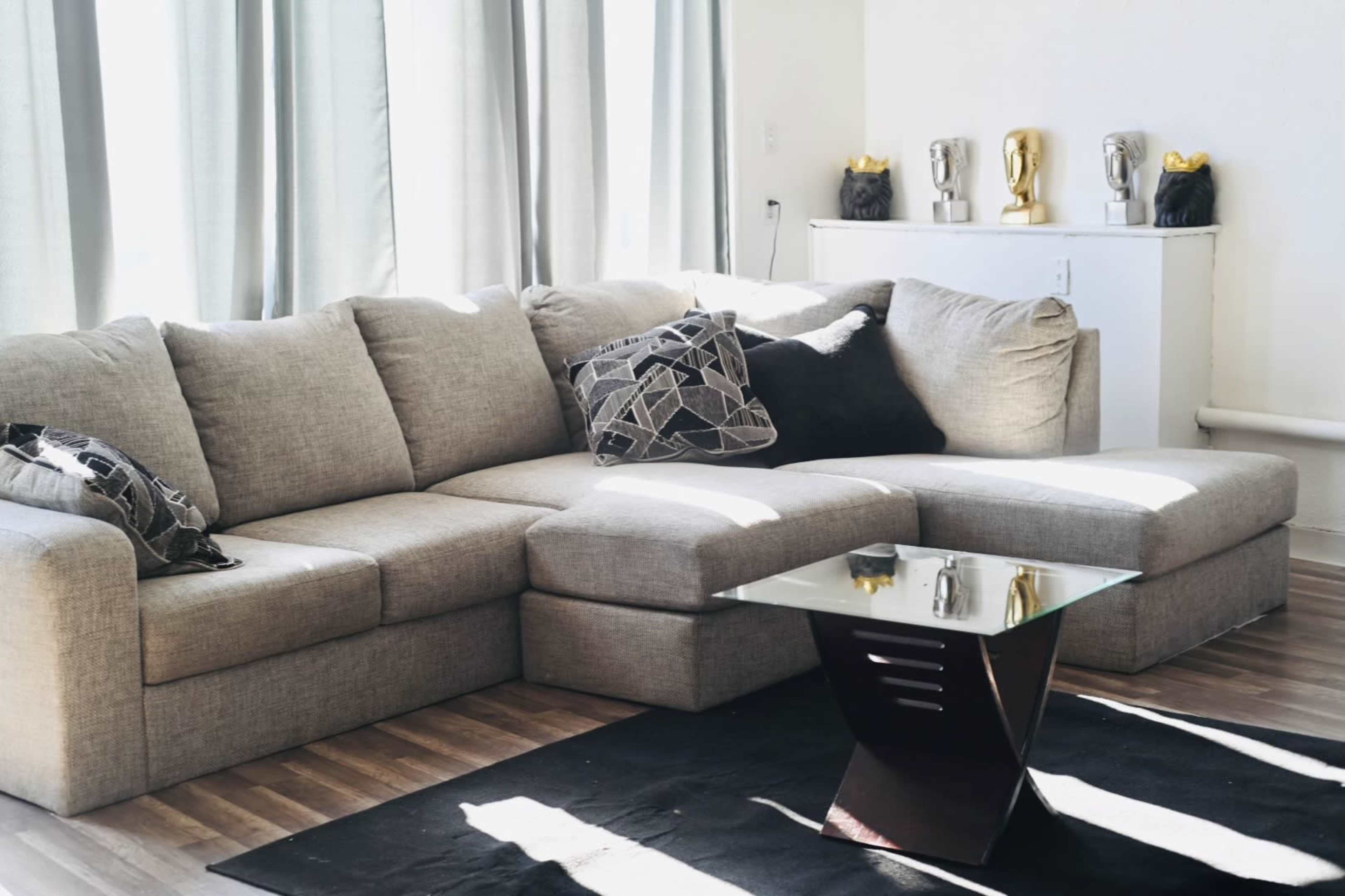A spacious living room features a gray sectional sofa with patterned pillows, a glass coffee table, and decorative items on a shelf.