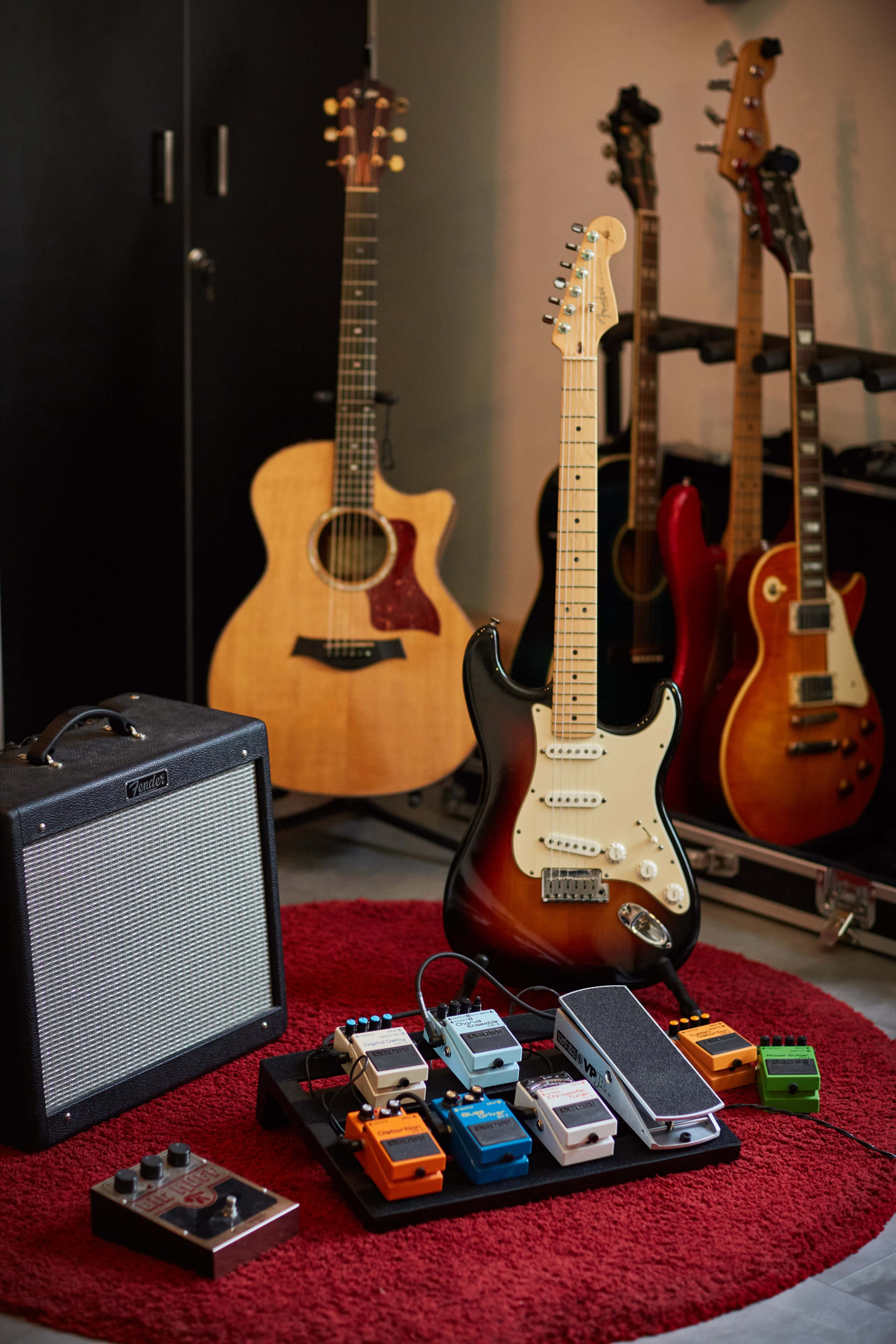 The image shows a collection of guitars and guitar pedals arranged on a red rug beside an amplifier in a music space.