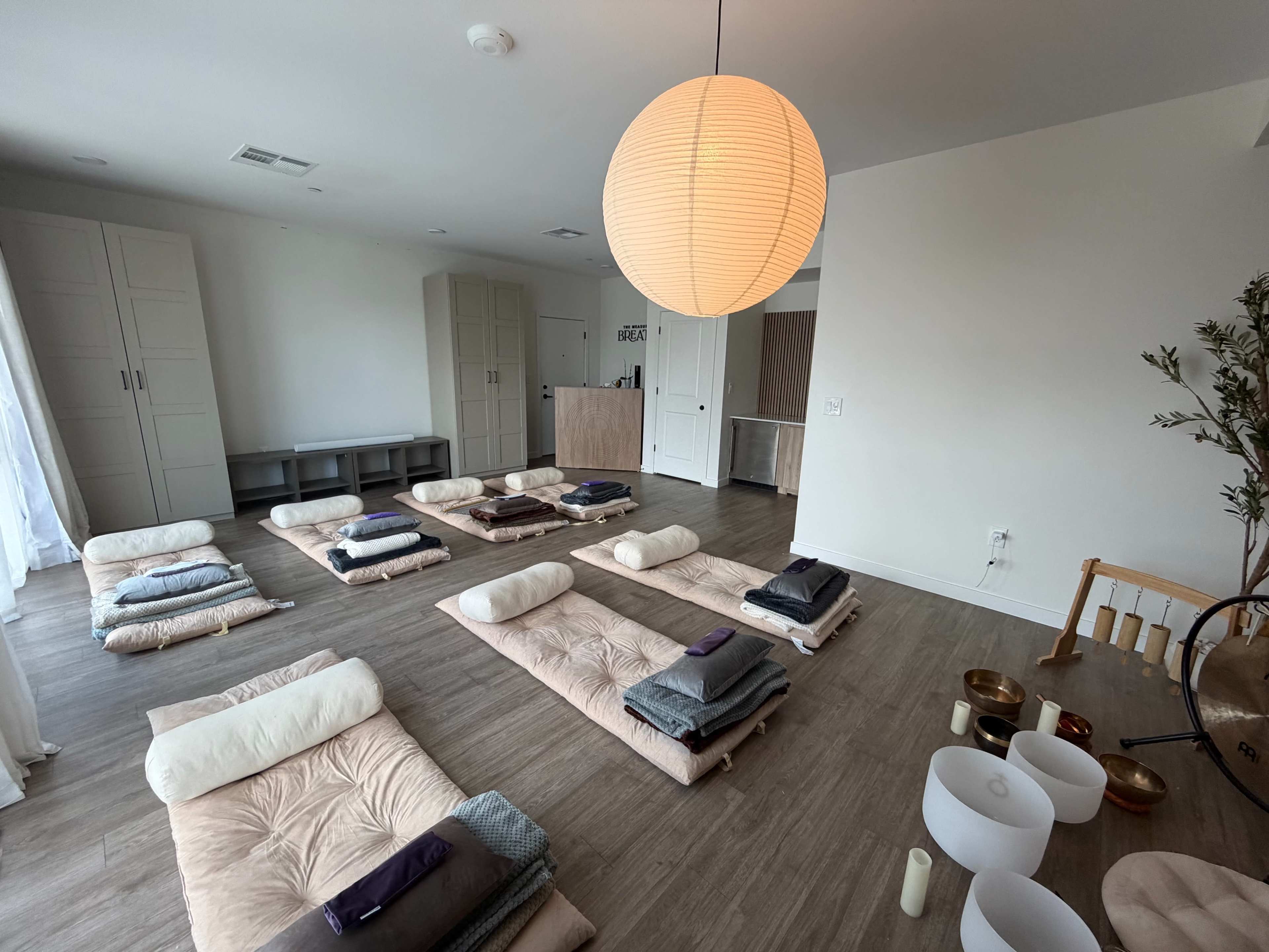 The image shows a serene room set up for a meditation session, with several mats and blankets arranged on the floor and a large pendant light hanging from the ceiling.