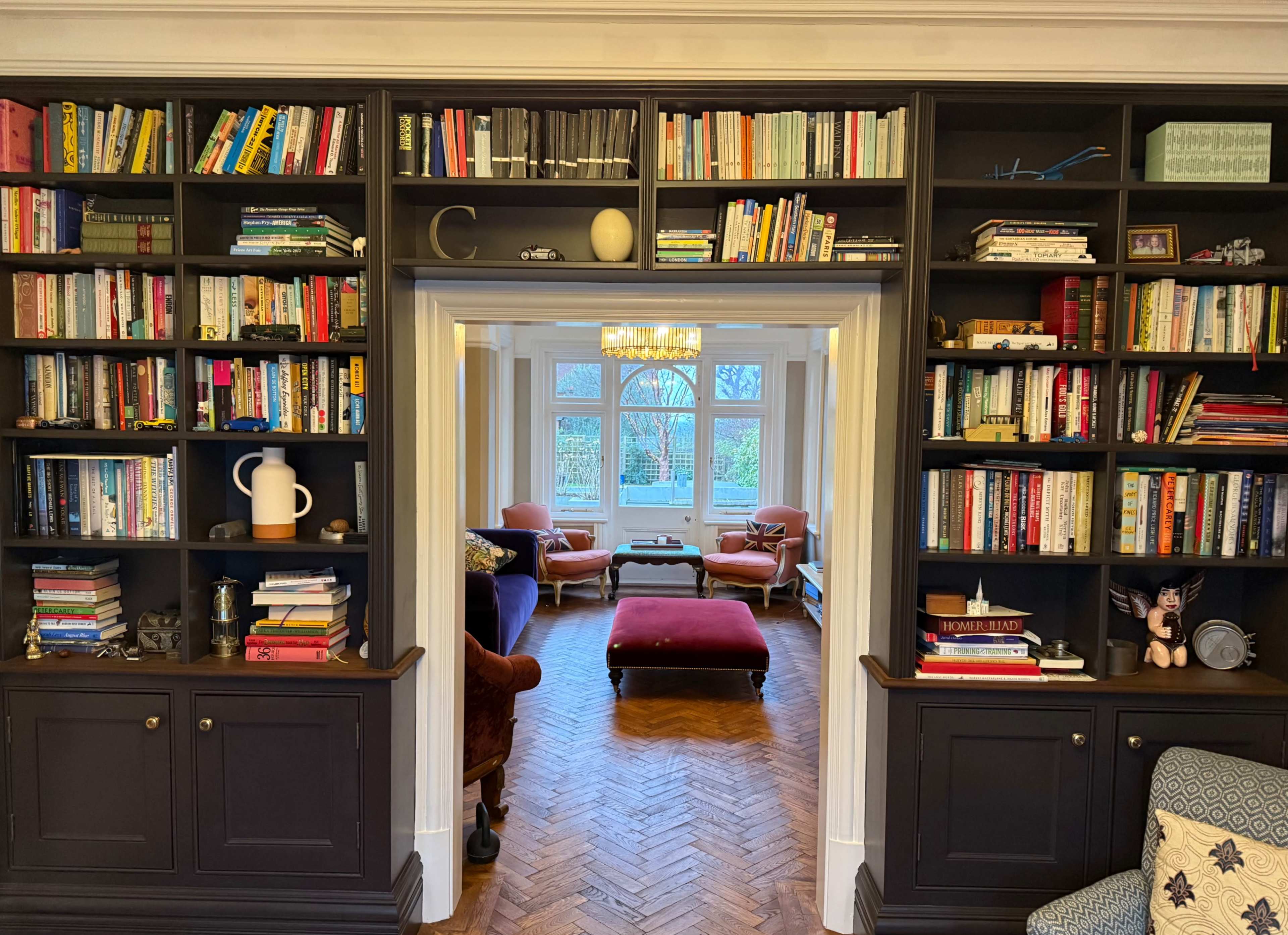 The image shows a room with a black bookshelf filled with books, leading to a light-filled seating area with a red ottoman and orange armchairs.
