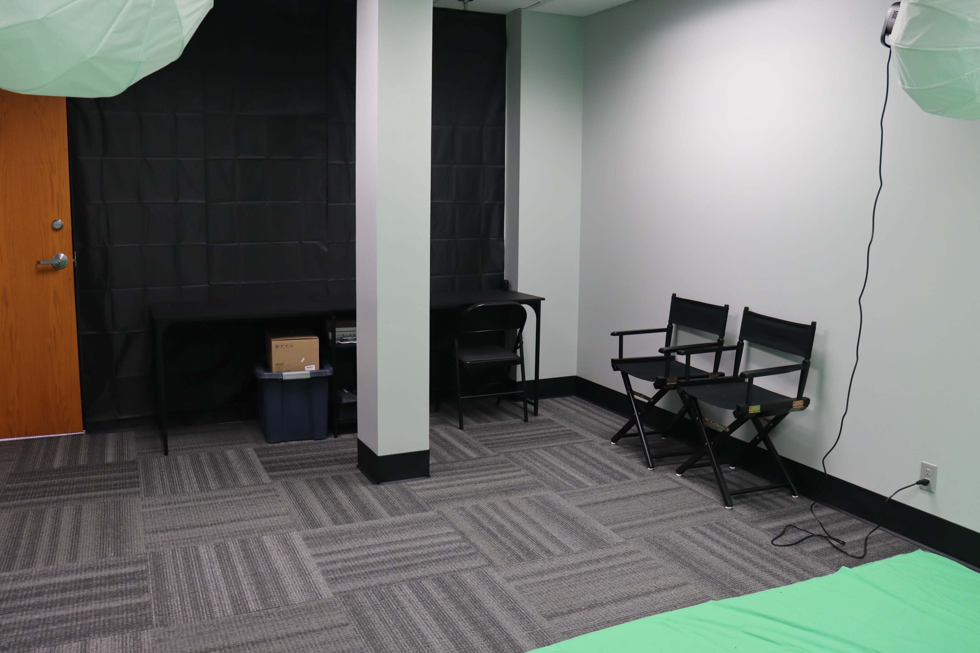 The image shows a room with black curtains, a green fabric on the floor, a desk with a chair, and two director's chairs.